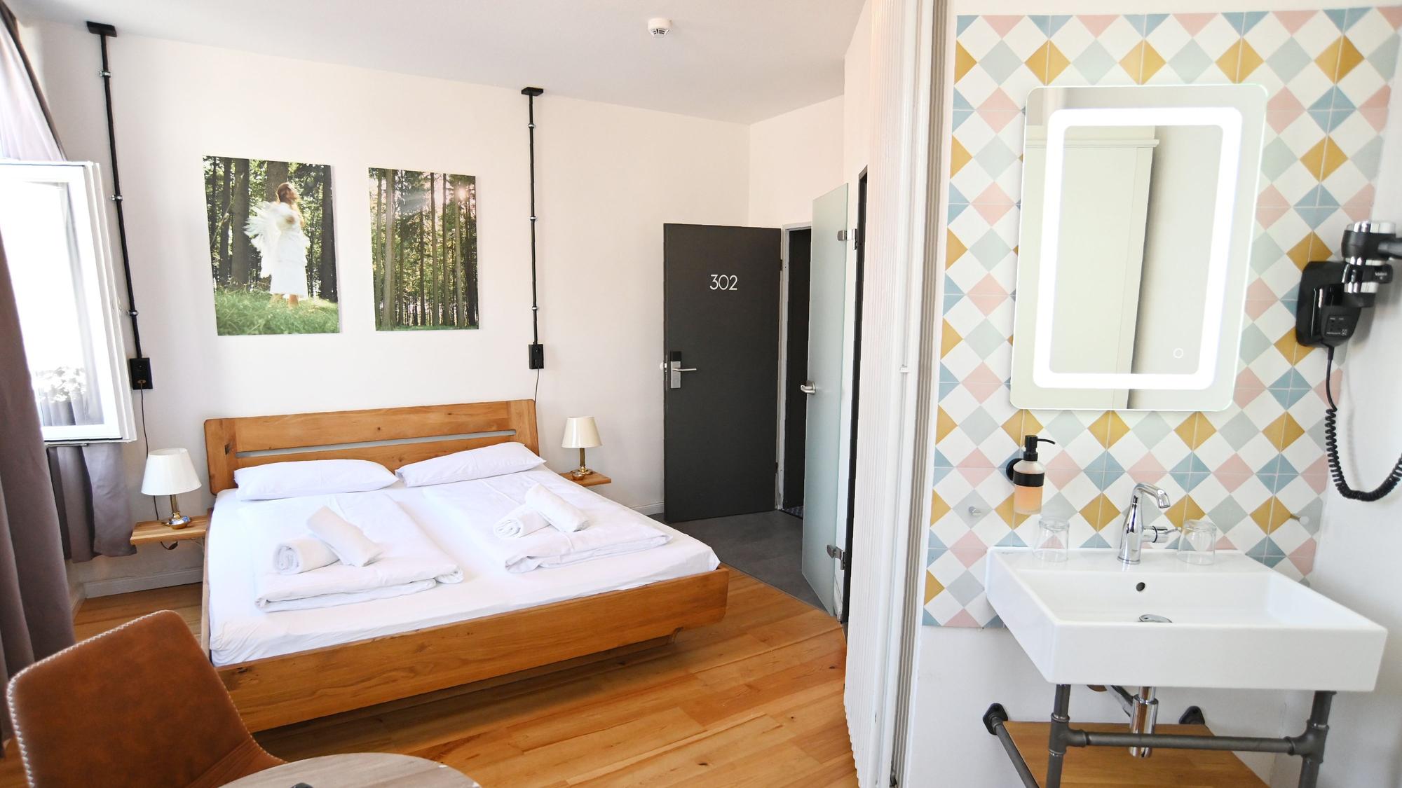 In einem hellen Zimmer steht ein Bett, in einer Nische ist ein Waschbecken, darunter ein Toiletten-Regal.