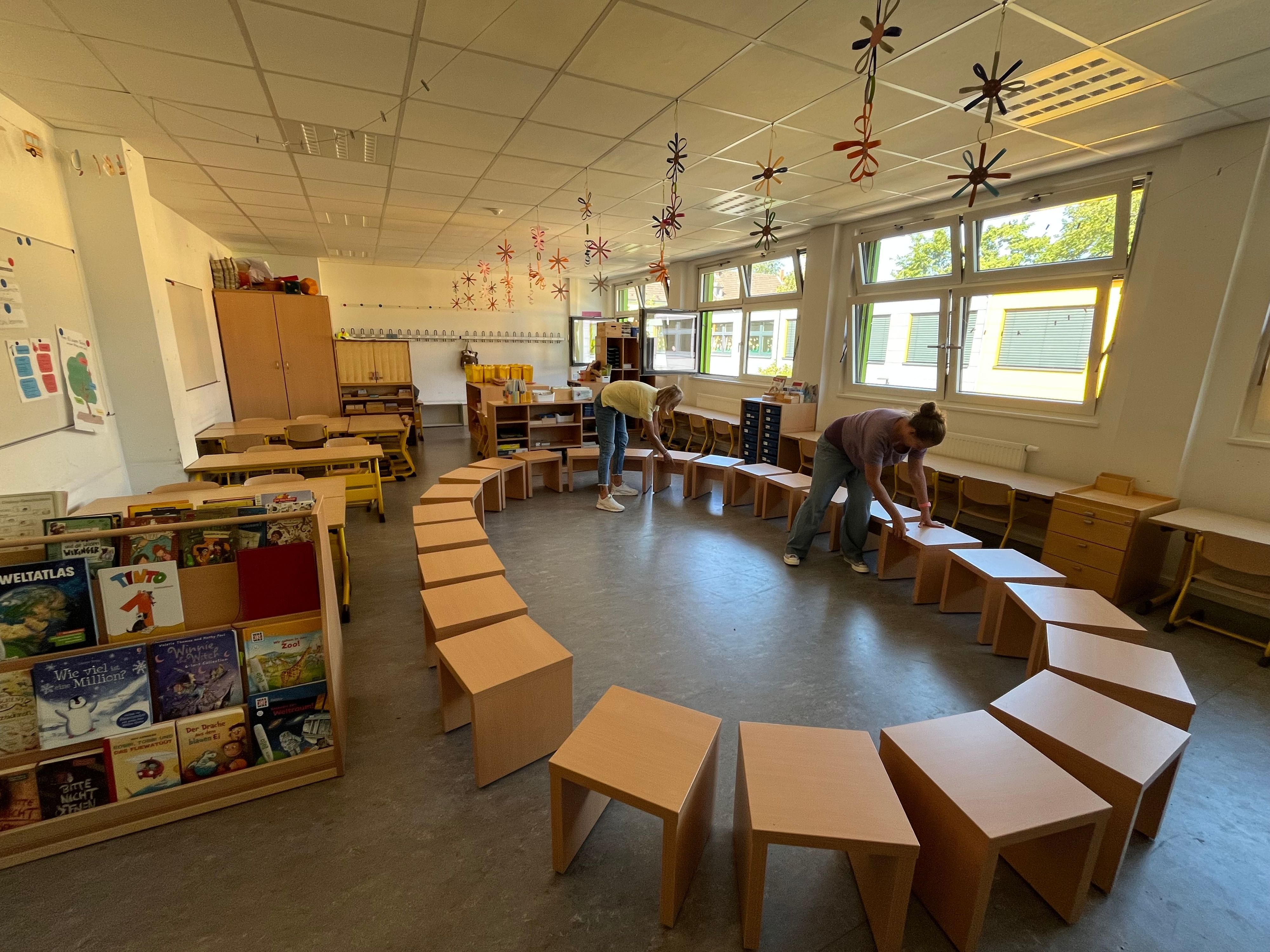 In einem Klassenraum stehen Bänke im Kreis, zwei Frauen schieben sie zurecht.