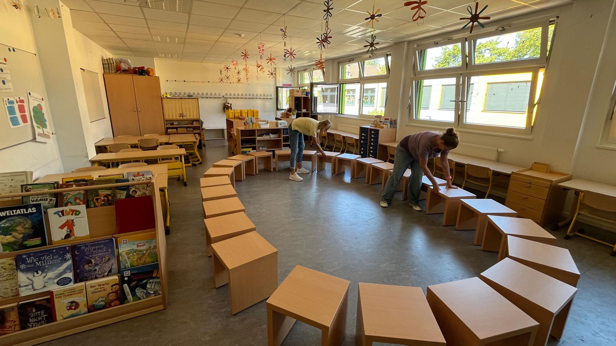 In einem Klassenraum stehen Bänke im Kreis, zwei Frauen schieben sie zurecht.