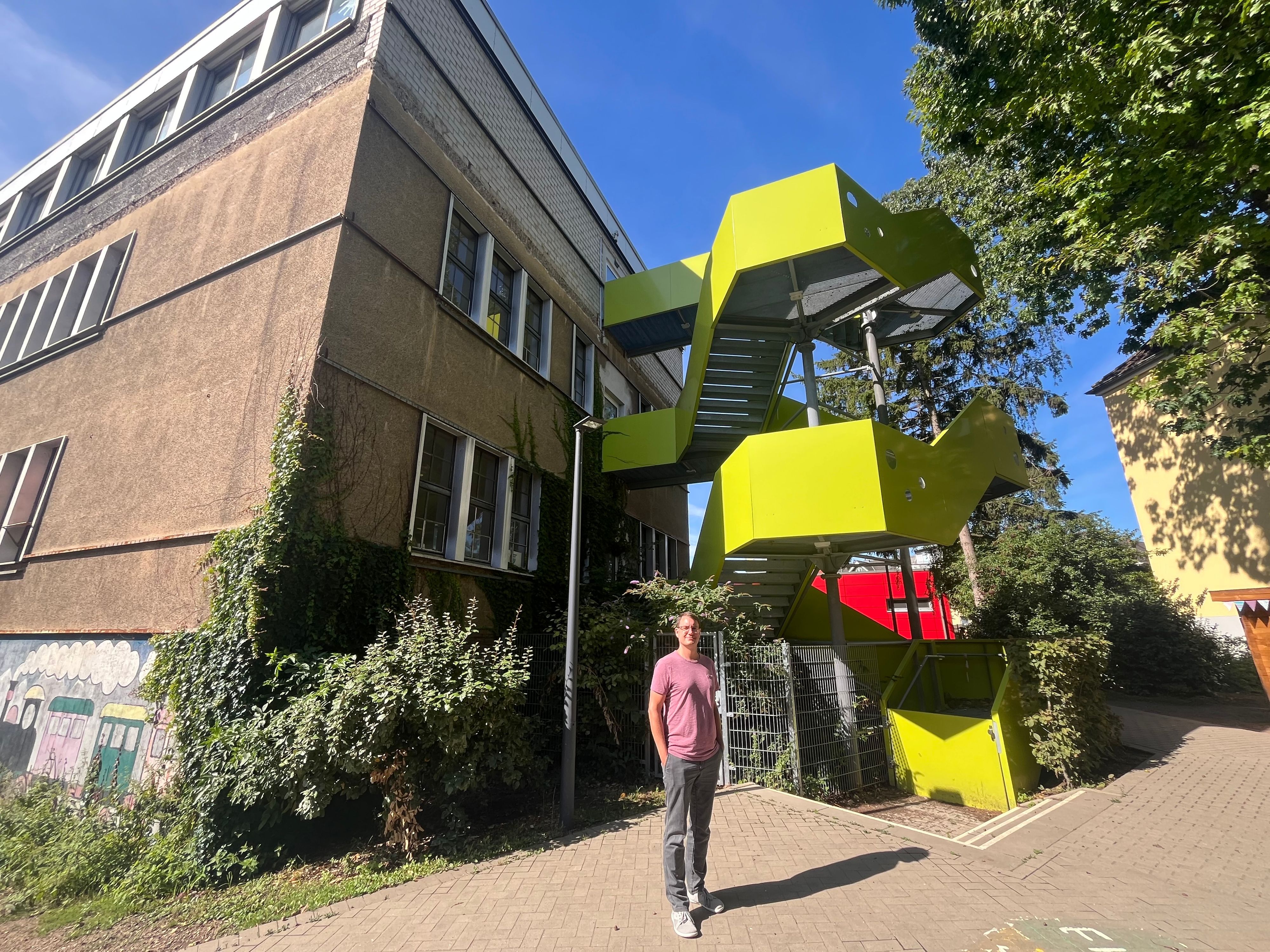 Ein Mann steht vor einem Schulgebäude  mit einer rotgelben Feuertreppe.