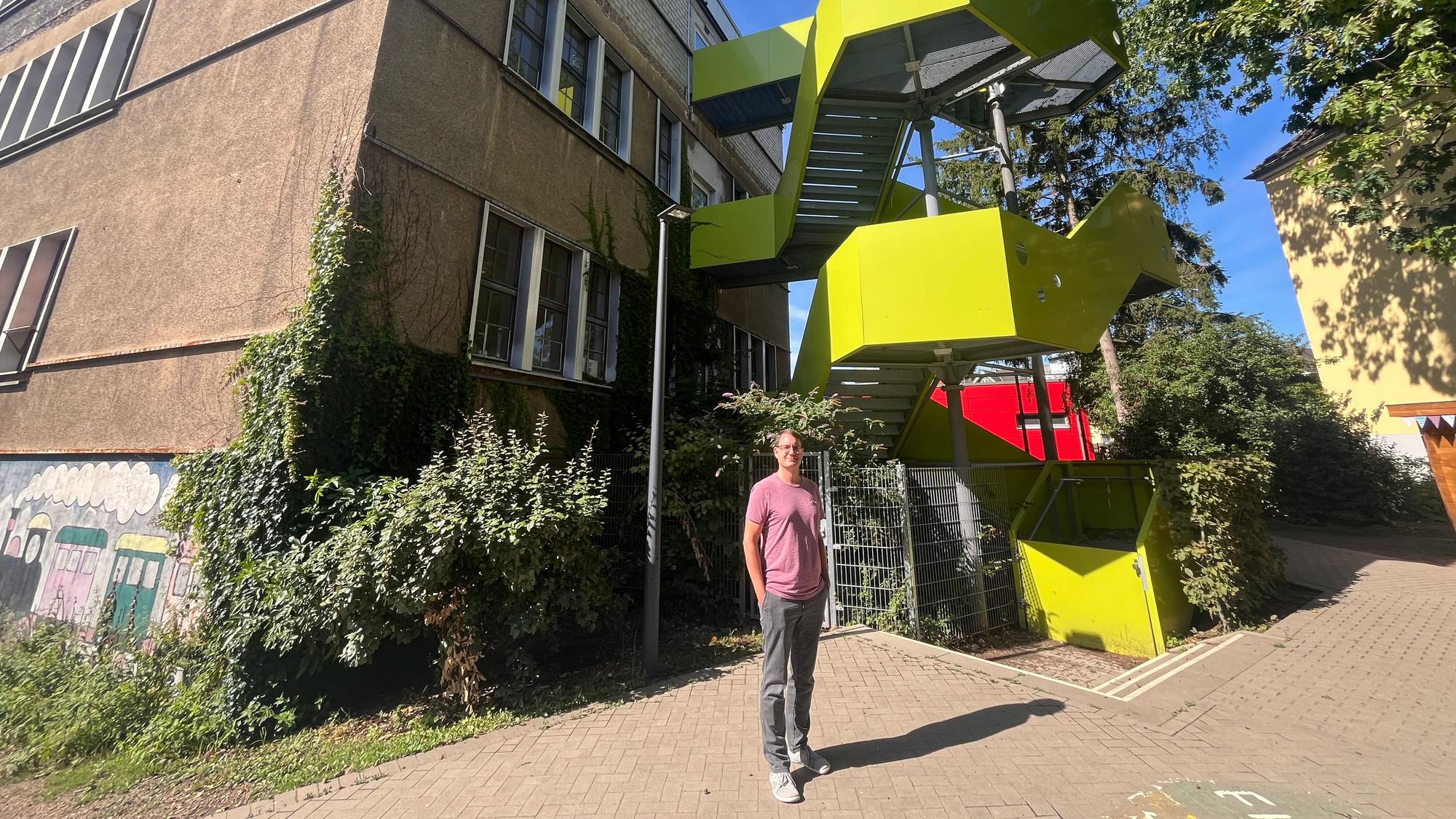 Ein Mann steht vor einem Schulgebäude mit einer rotgelben Feuertreppe.