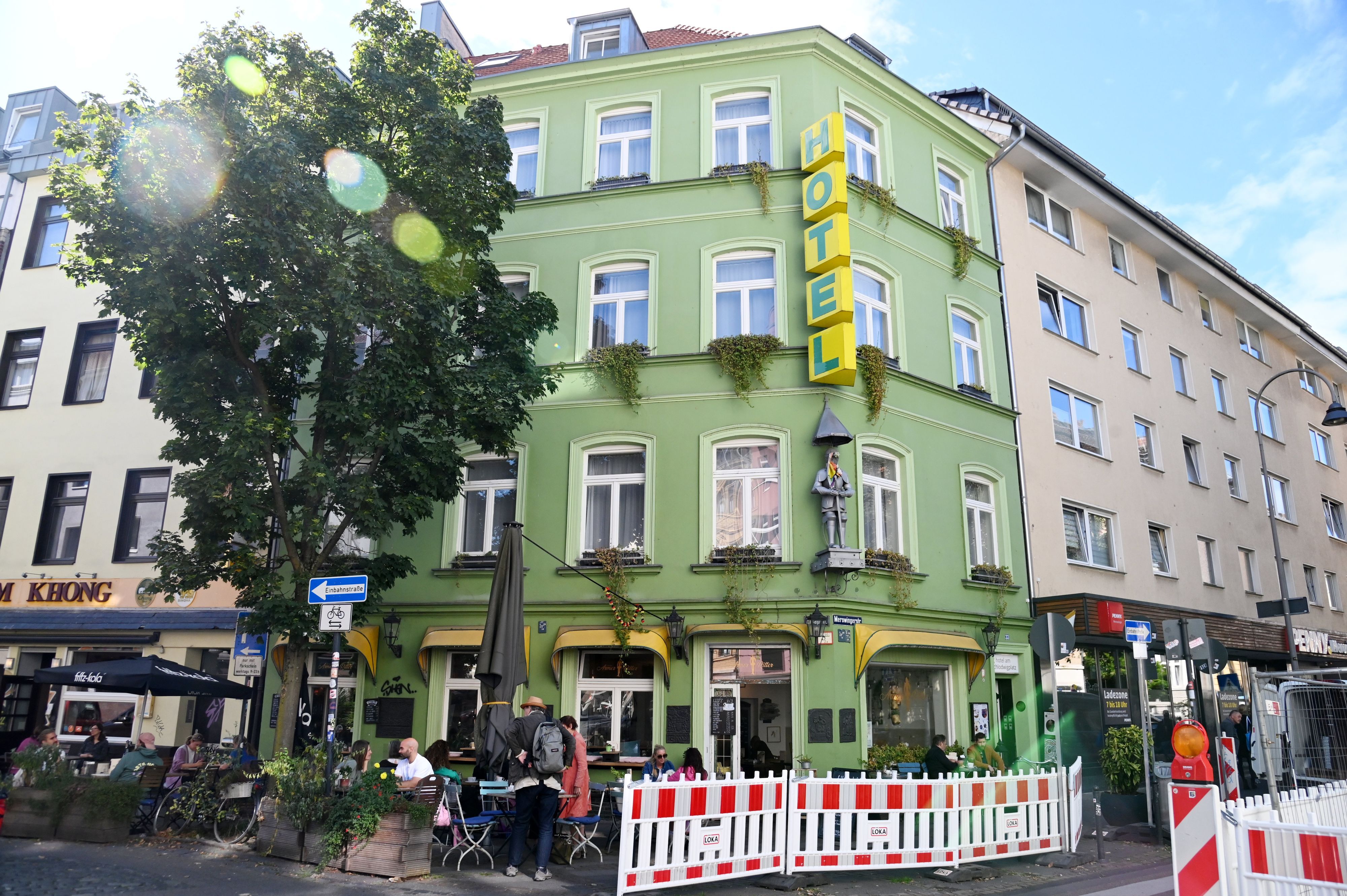 Hinter rot-weißen Absperrvorrichtungen ist ein grünes Eckgebäude zu sehen, vor dem Haus sitzen Menschen an Café-Tischen.