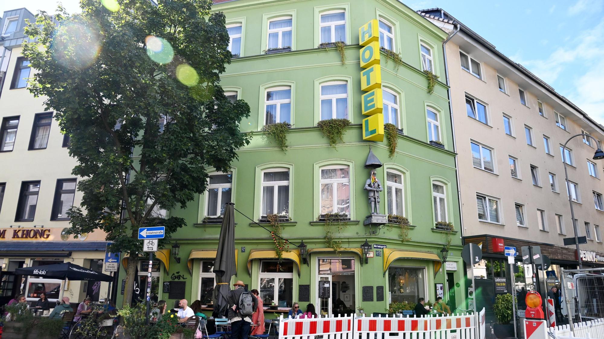 Hinter rot-weißen Absperrvorrichtungen ist ein grünes Eckgebäude zu sehen, vor dem Haus sitzen Menschen an Café-Tischen.