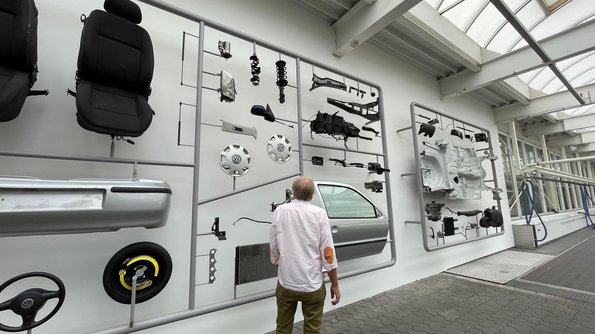 Mann steht in einer Halle, an den Wänden hängen große Tafeln, auf denen Autoteile zu sehen sind.