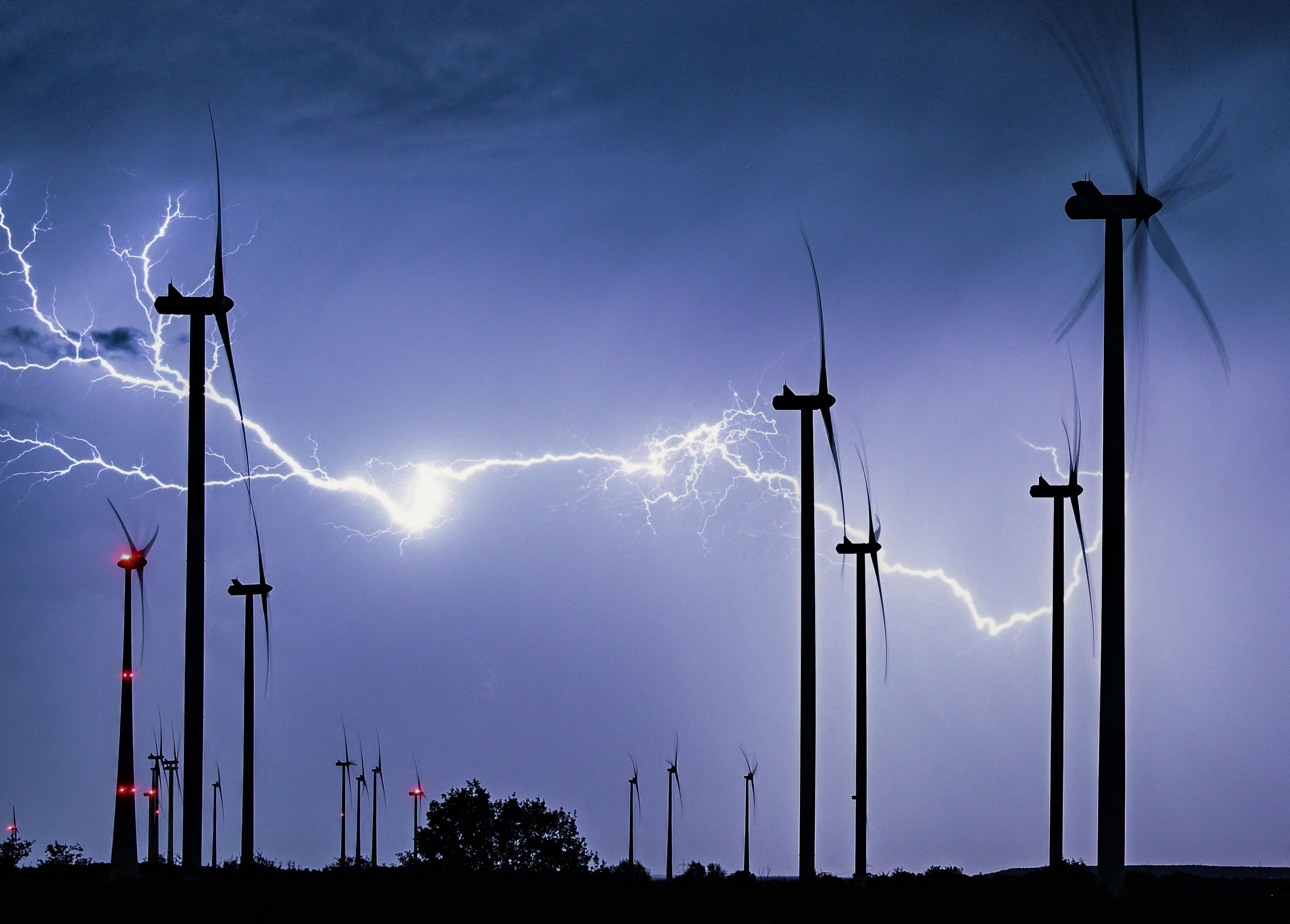 Das Symbolfoto zeigt einen mächtigen verzweigten Gewitterblitz, der die nächtliche Landschaft eines Windparks erleuchtet.