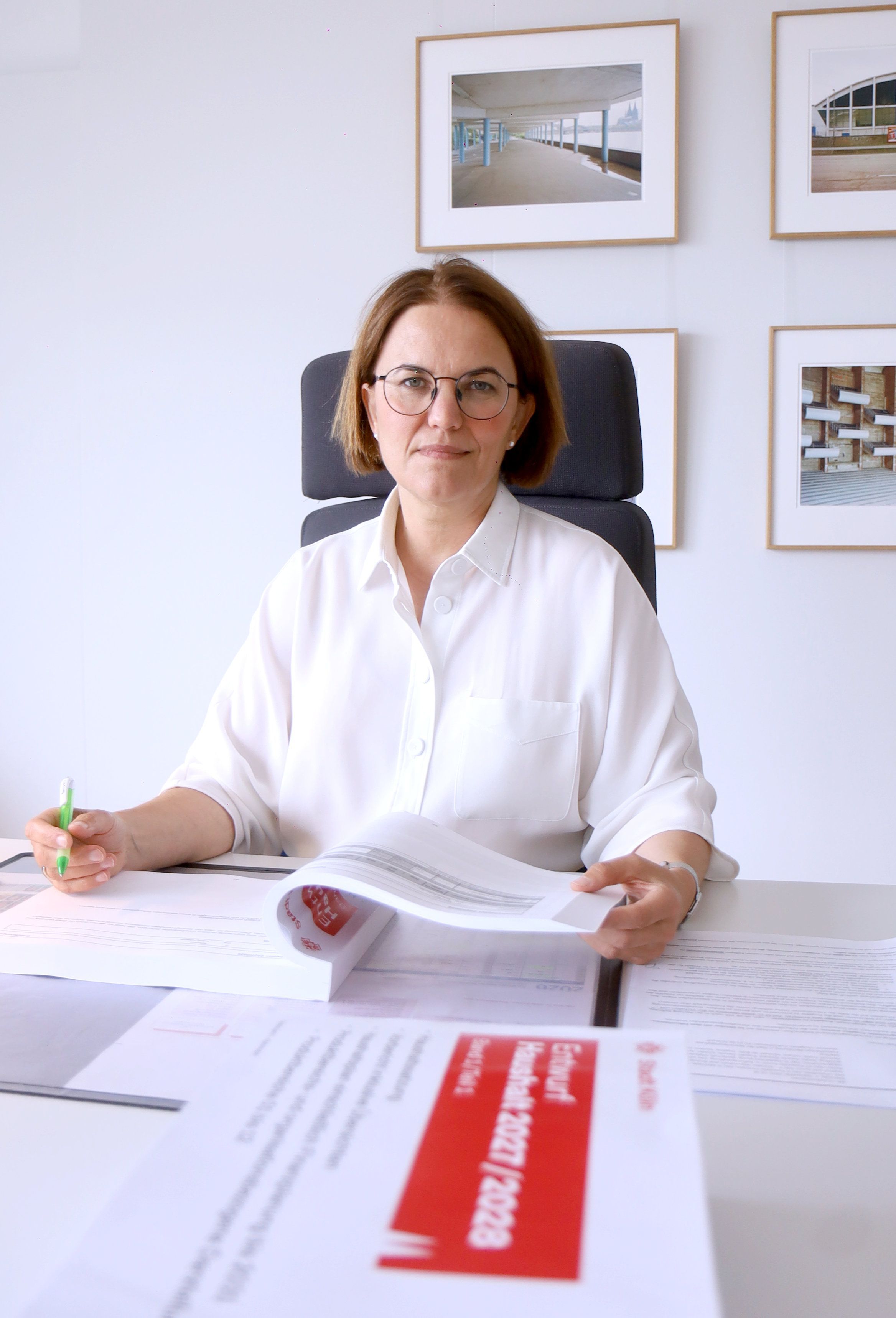 Eine Frau mit brünettem Pagenkopf und Brille sitzt an einem Schreibtisch. Hinter ihr hängen Bilder an der Wand.