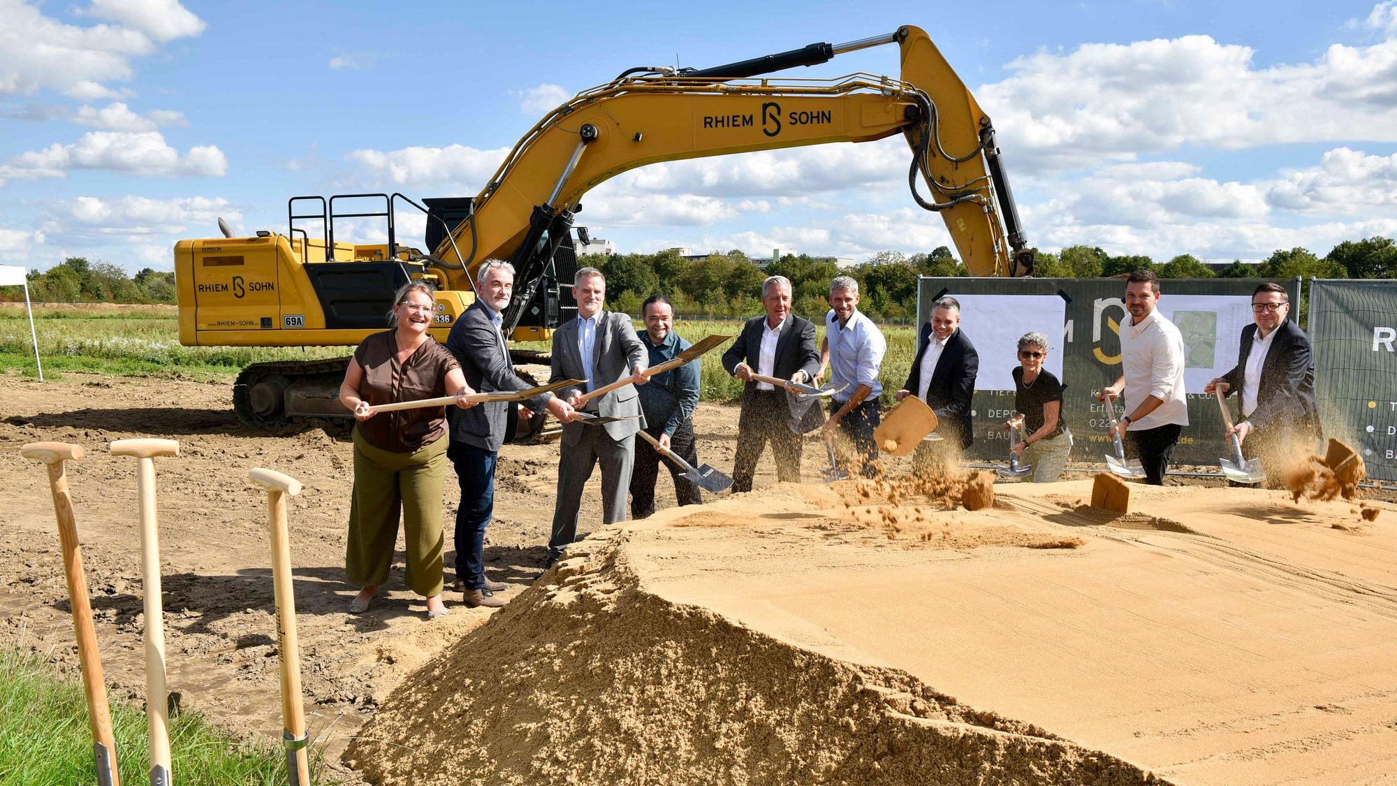 Das Foto zeigt den symbolischen ersten Spatenstich mit Bagger im Hintergrund.
