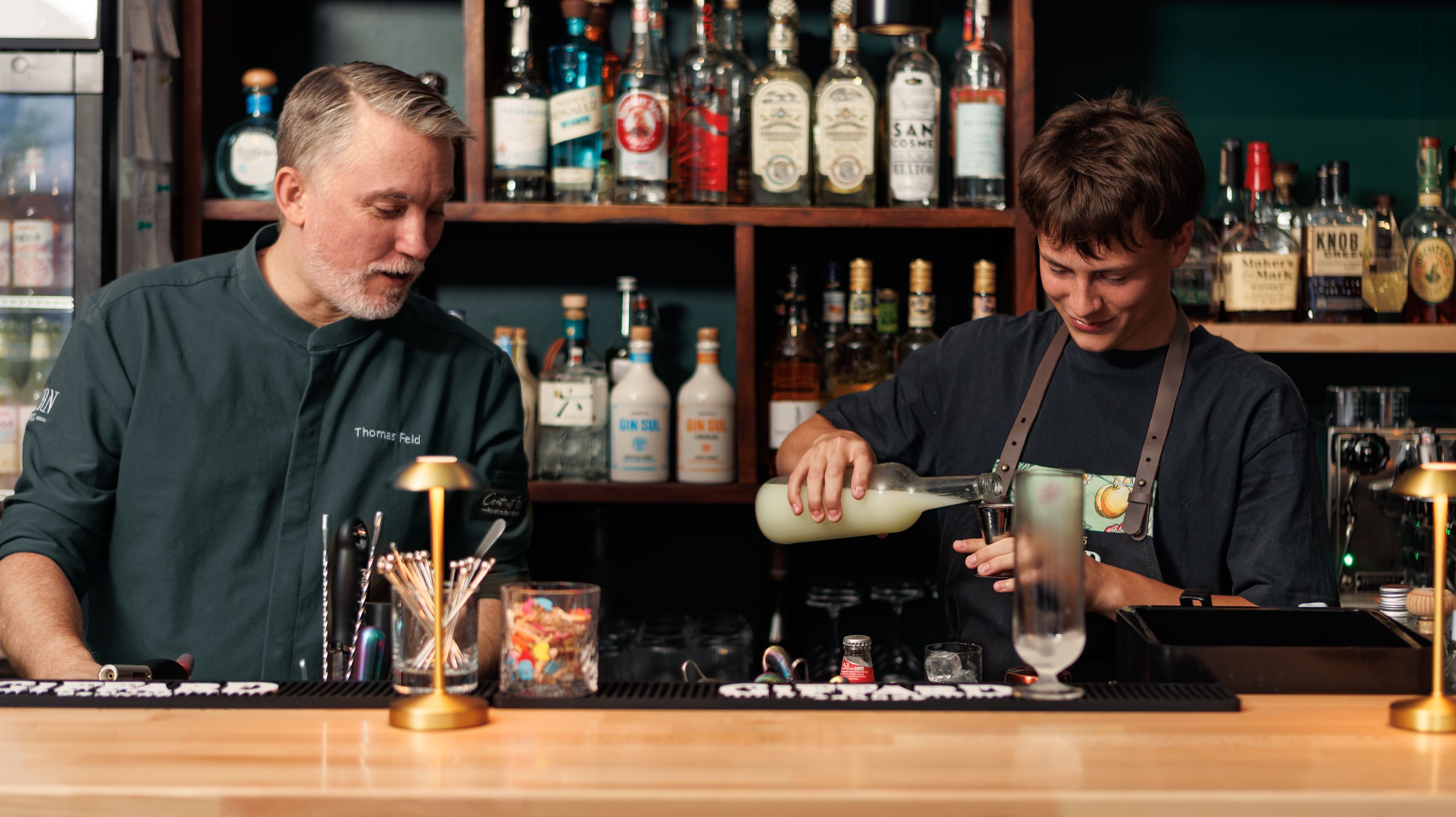 Zwei Männer in dunkler Bartender-Kleidung stehen hinter einer Theke.