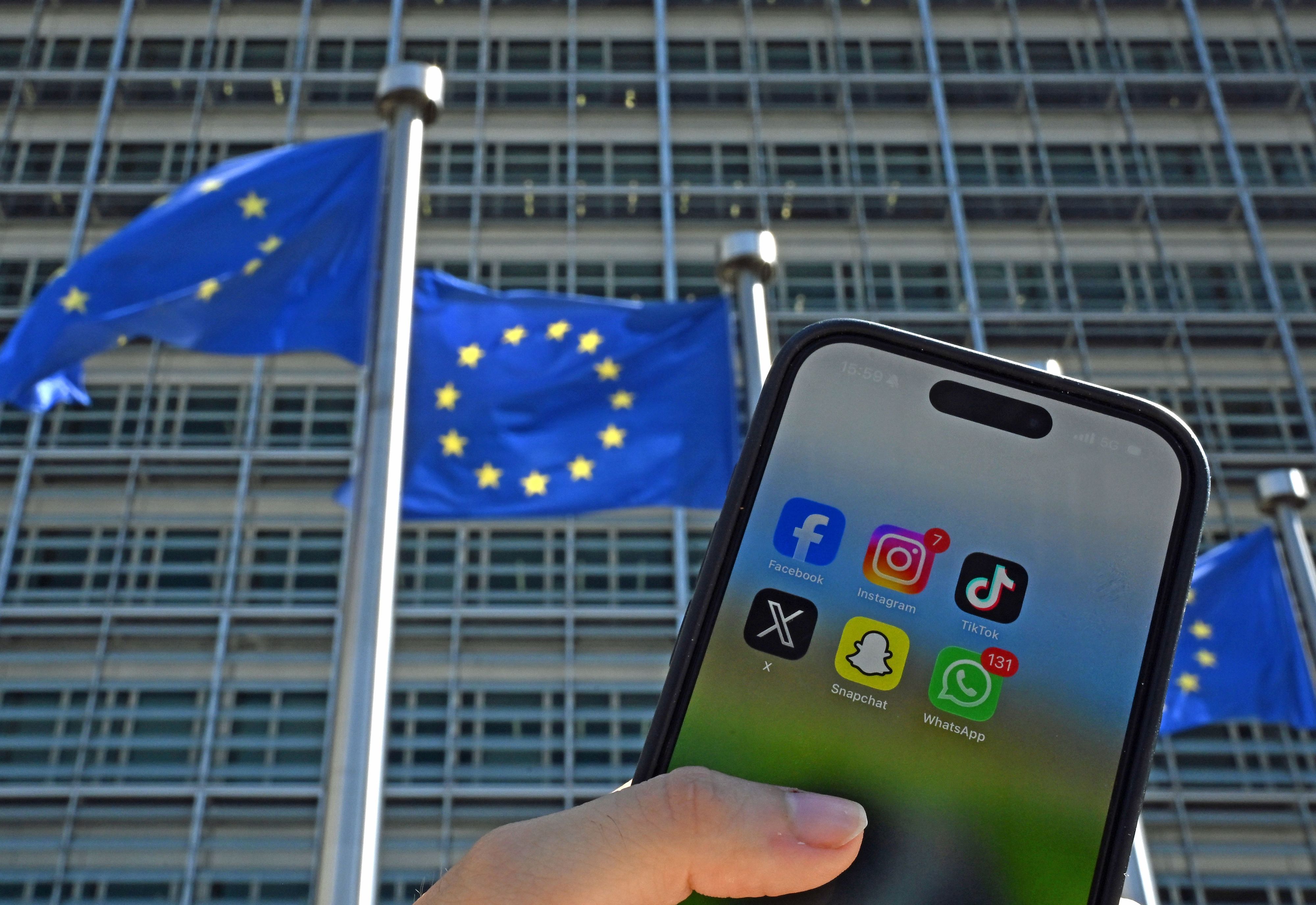 Eine Person hält ein Smartphone mit den Logos der Social-Media-Apps Facebook, Instagram, TikTok, X, Snapchat und WhatsApp vor Fahnen der Europäischen Union und dem Gebäude der Europäischen Kommission. Die EU-Kommission will den Zugang zu Social Media für Kinder einschränken.