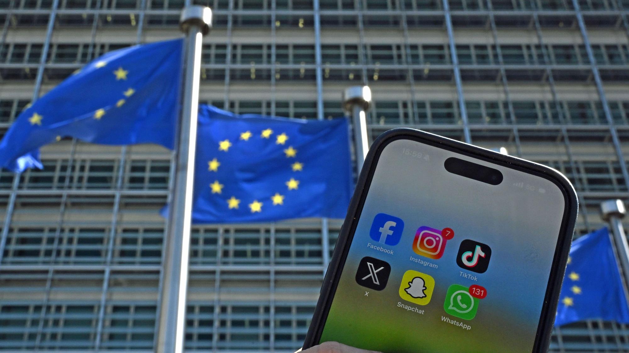 Eine Person hält ein Smartphone mit den Logos der Social-Media-Apps Facebook, Instagram, TikTok, X, Snapchat und WhatsApp vor Fahnen der Europäischen Union und dem Gebäude der Europäischen Kommission. Die EU-Kommission will den Zugang zu Social Media für Kinder einschränken.