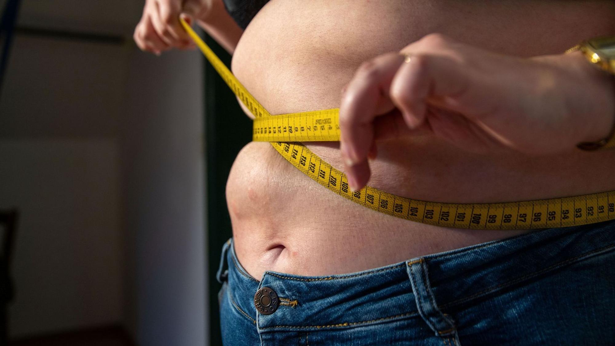 Eine Frau mit Übergewicht hält ein Maßband um ihren Bauch.