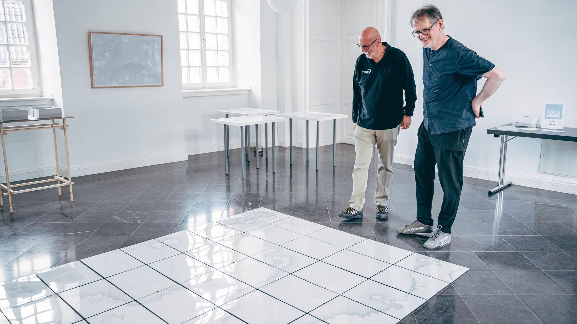 Auf dem Bild sind zwei Männer vor einem Bodenkunstwerk zu sehen.