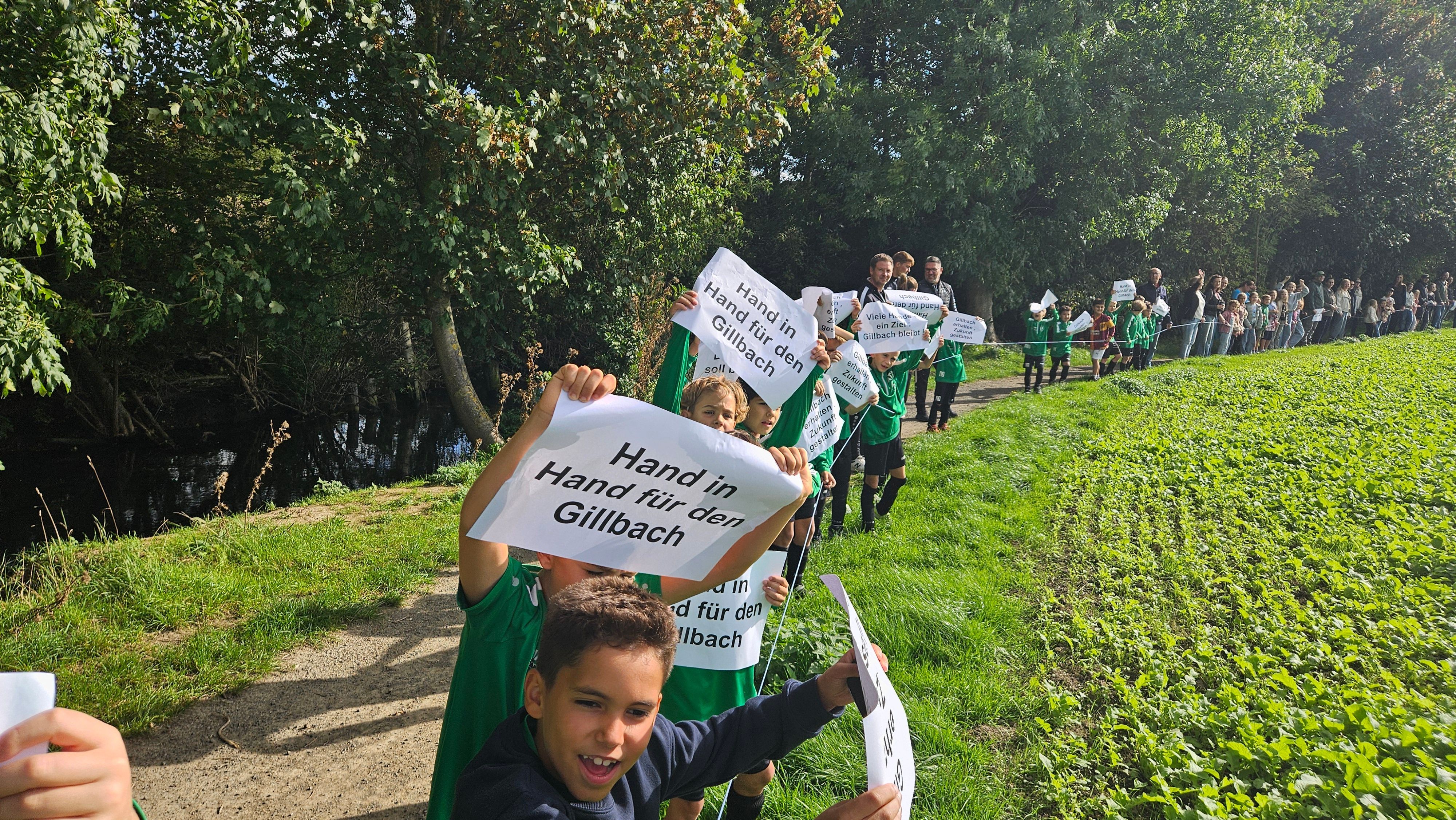 Zu sehen sind Kinder und Erwachsene entlang eines Bachs. Sie tragen Zettel, mit denen sie für den Erhalt des Gillbachs eintreten.