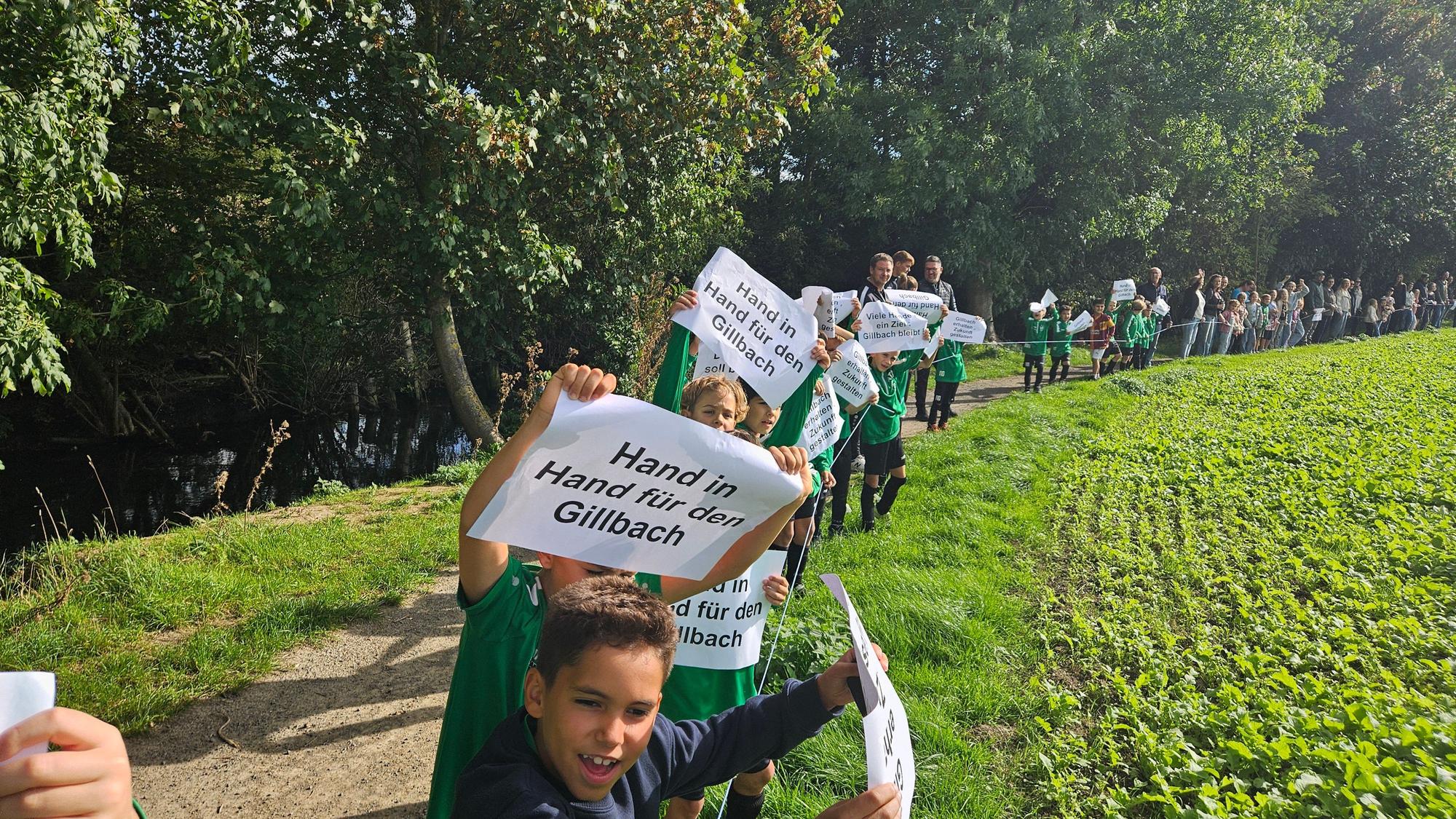 Zu sehen sind Kinder und Erwachsene entlang eines Bachs. Sie tragen Zettel, mit denen sie für den Erhalt des Gillbachs eintreten.