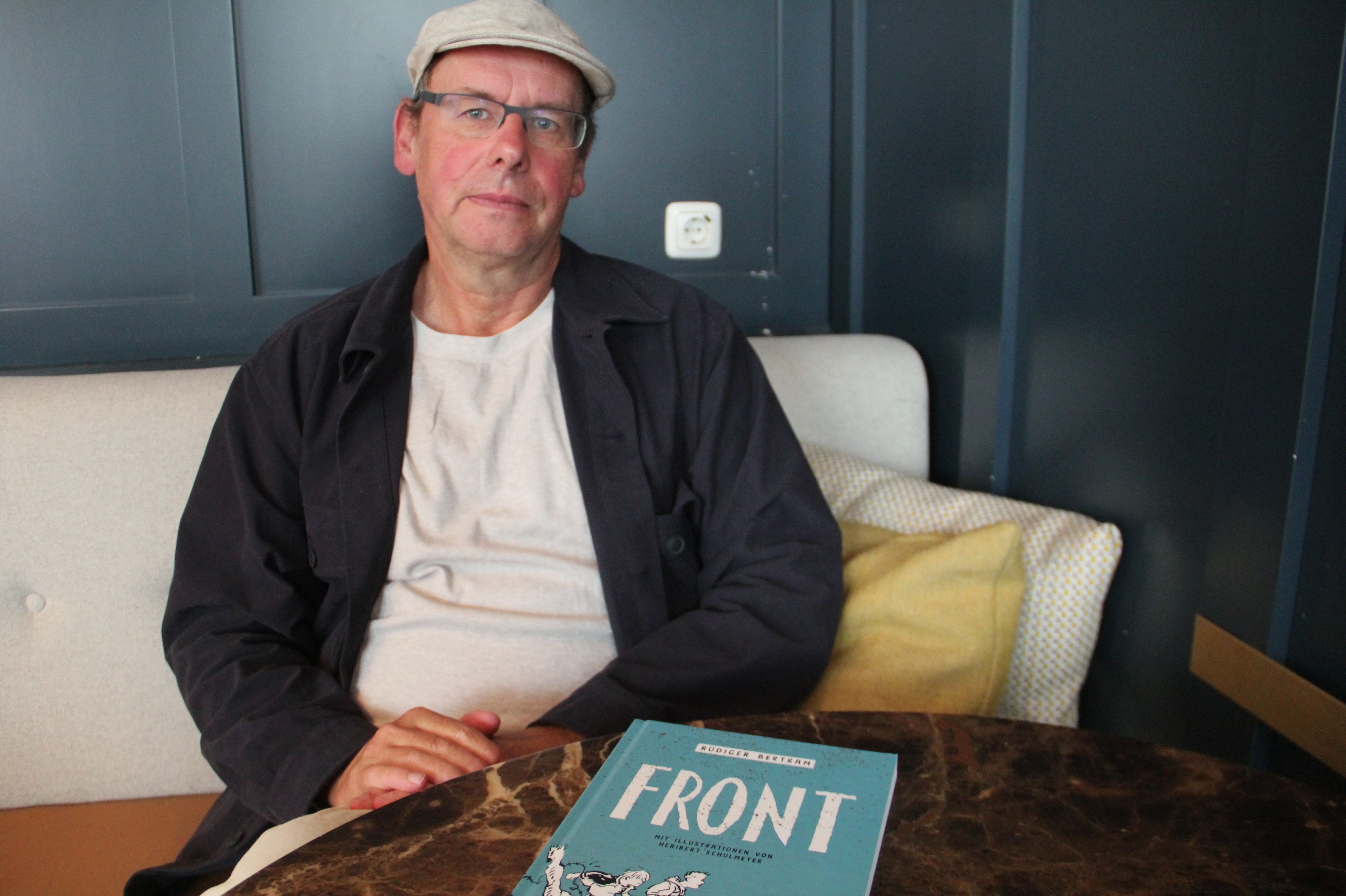 Ein Mann mit Kappe und Brille sitzt auf einer Bank. Vor ihm auf dem Tisch liegt sein neues Buch „Front“.