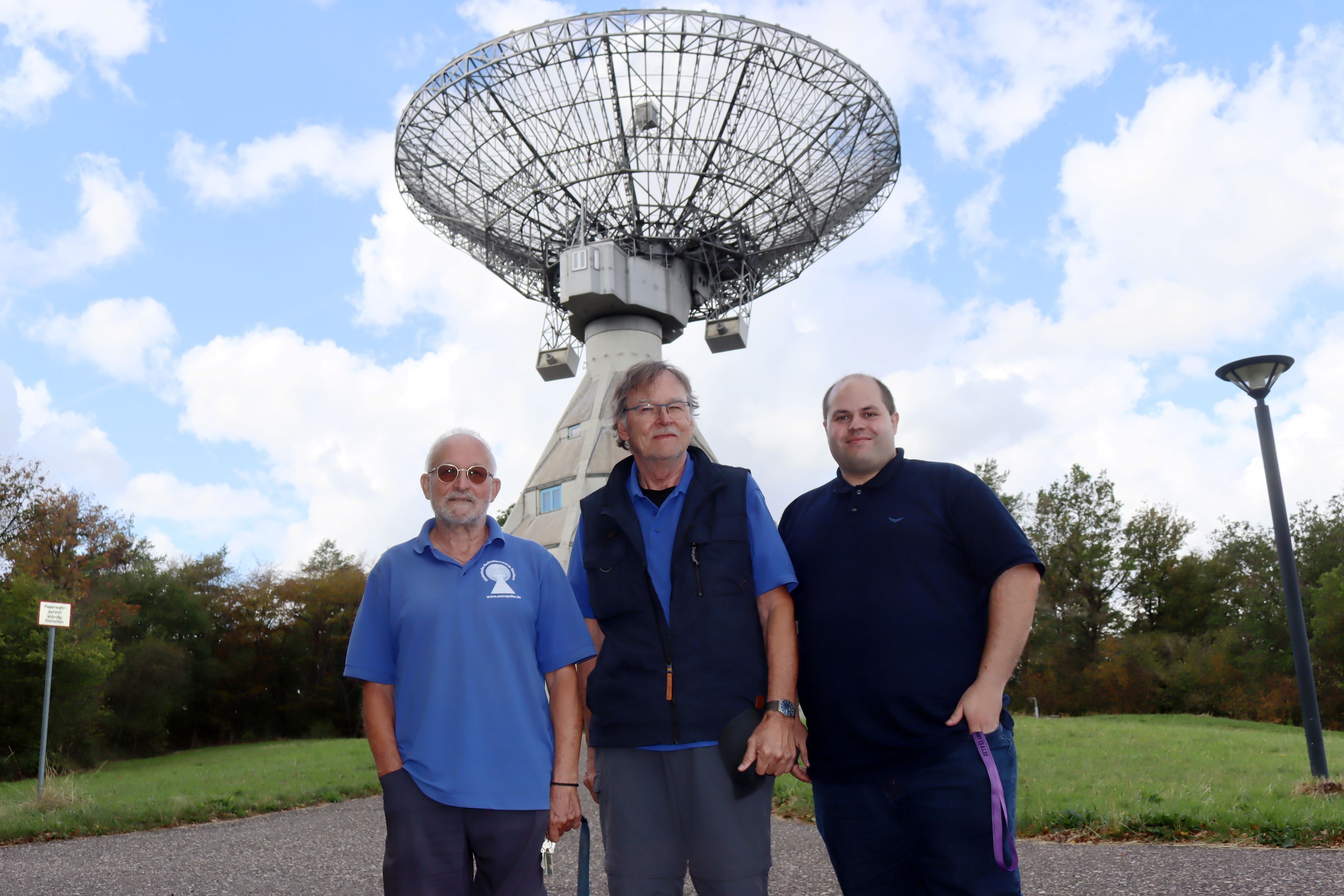 Drei Männer stehen nebeneinander, im Hintergrund thront der 25-Meter-Spiegel des Astropeilers.