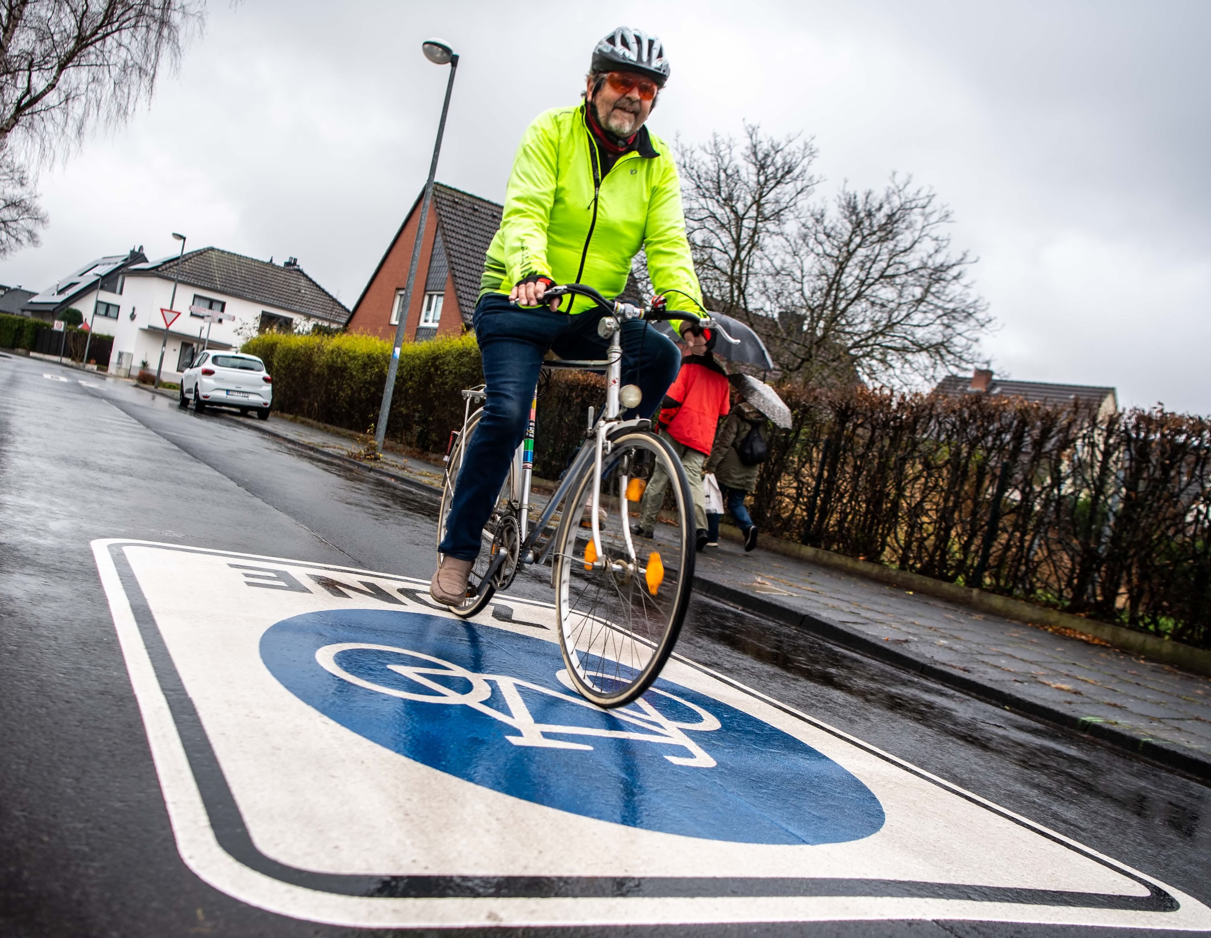 Das Bild zeigt einen Radfahrer mit Helm und leuchtend gelber Jacke in einem Wohngebiet.