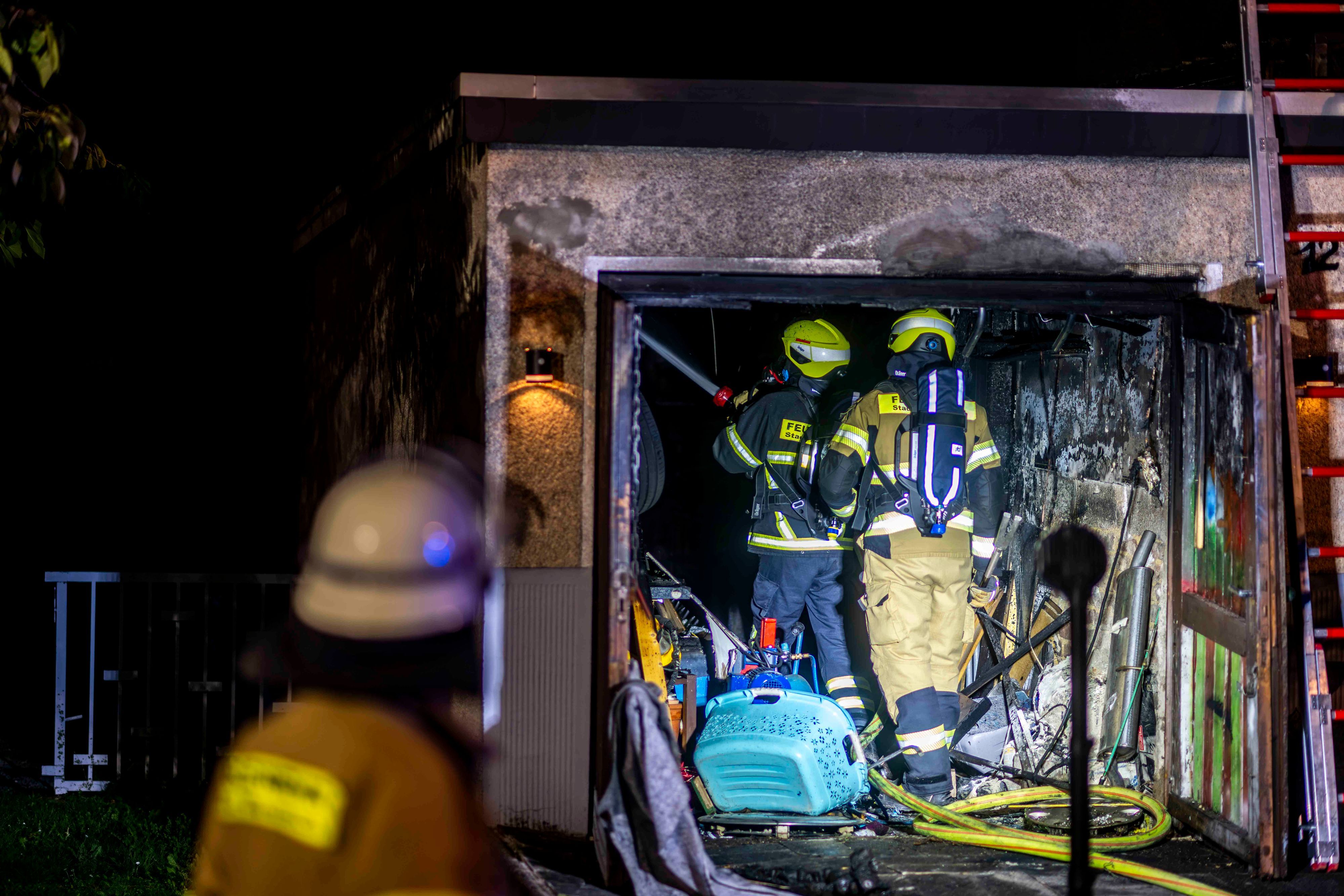 Zwei Feuerwehrleute stehen in der verrußten Garage, links ist im Vordergrund ein Feuerwehrmann von hinten unscharf zu sehen.