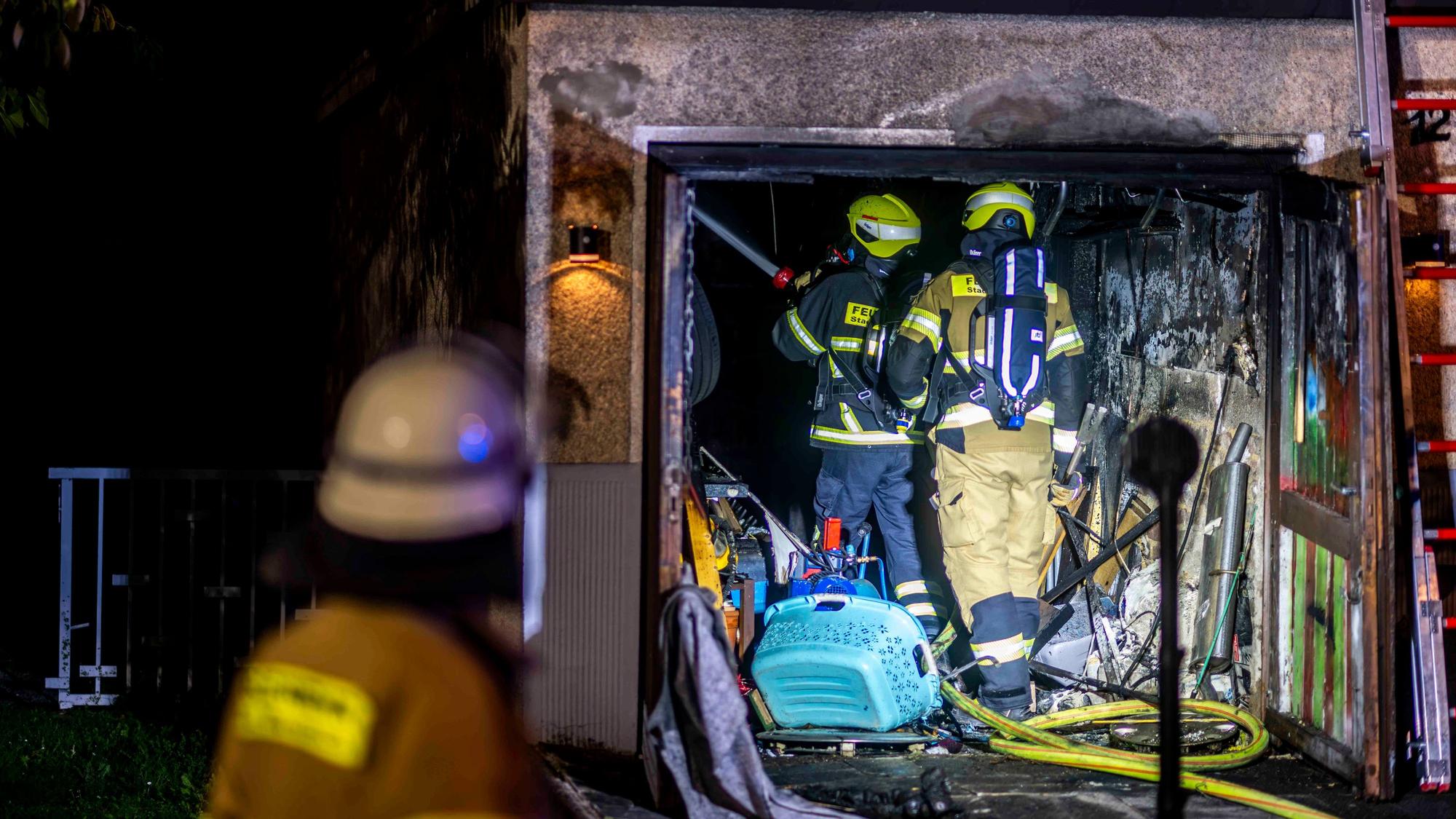 Zwei Feuerwehrleute stehen in der verrußten Garage, links ist im Vordergrund ein Feuerwehrmann von hinten unscharf zu sehen.