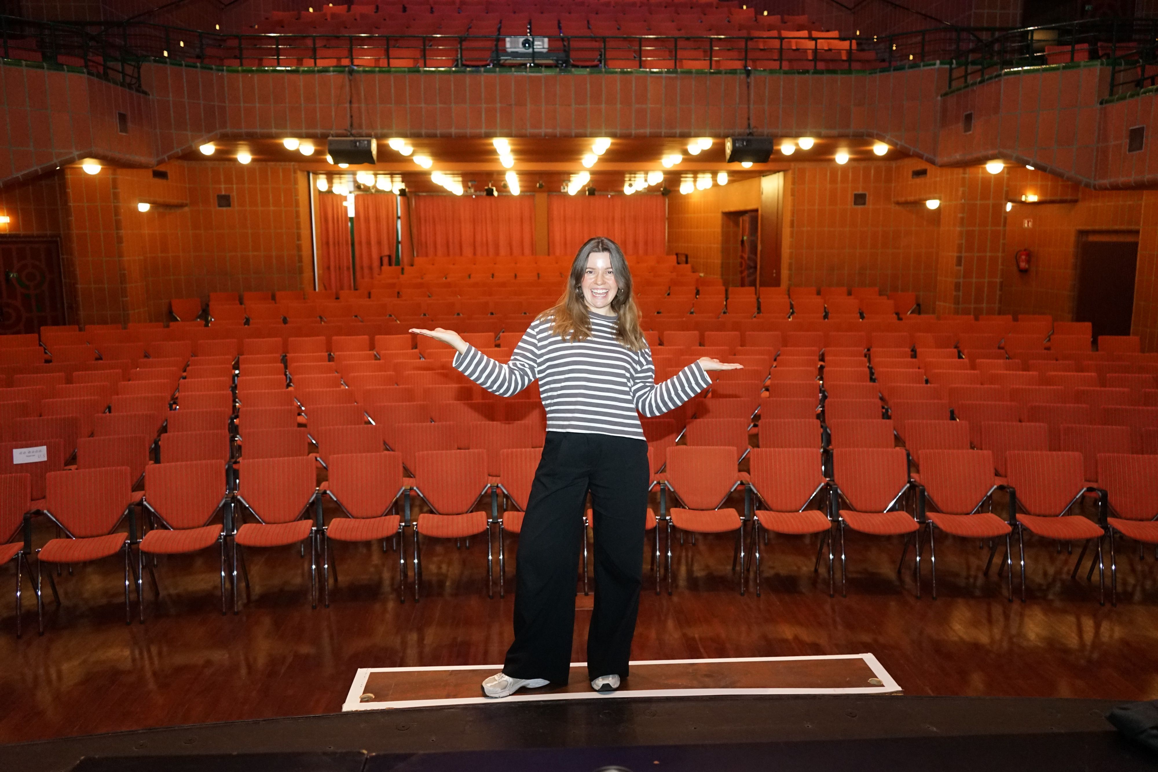 Eine Frau steht mit ausgebreiteten Armen auf einer Theaterbühne, hinter ihr rote Stuhlreihen.