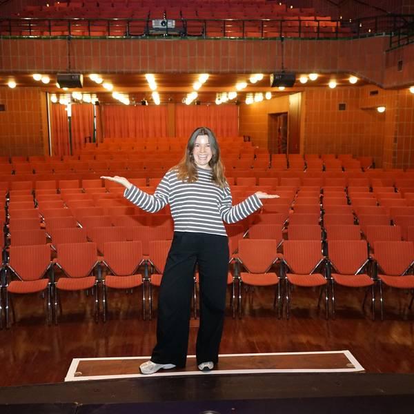 Eine Frau steht mit ausgebreiteten Armen auf einer Theaterbühne, hinter ihr rote Stuhlreihen.