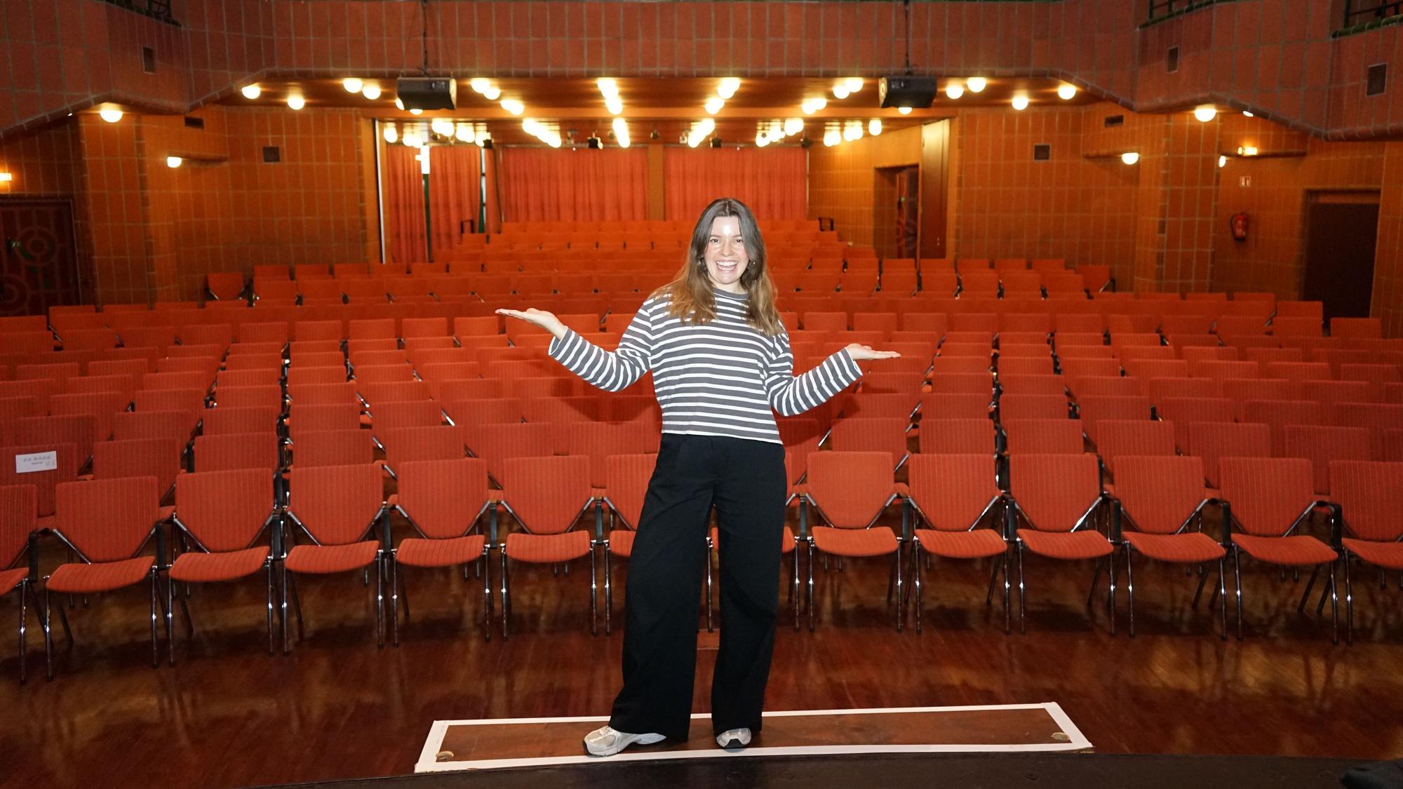 Eine Frau steht mit ausgebreiteten Armen auf einer Theaterbühne, hinter ihr rote Stuhlreihen.
