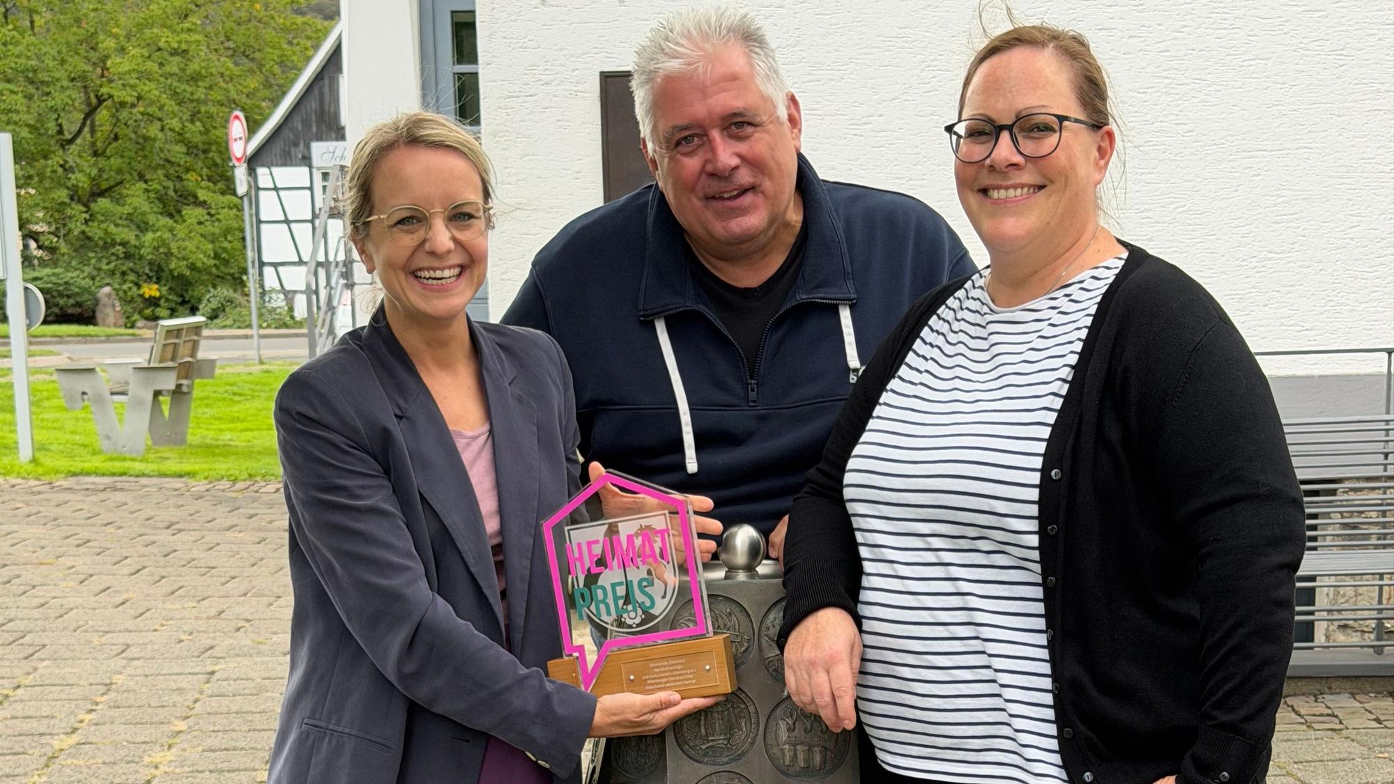 Zwei Frauen und ein Mann lachen in die Kamera, eine Frau hält den Heimatpreis in den Händen.