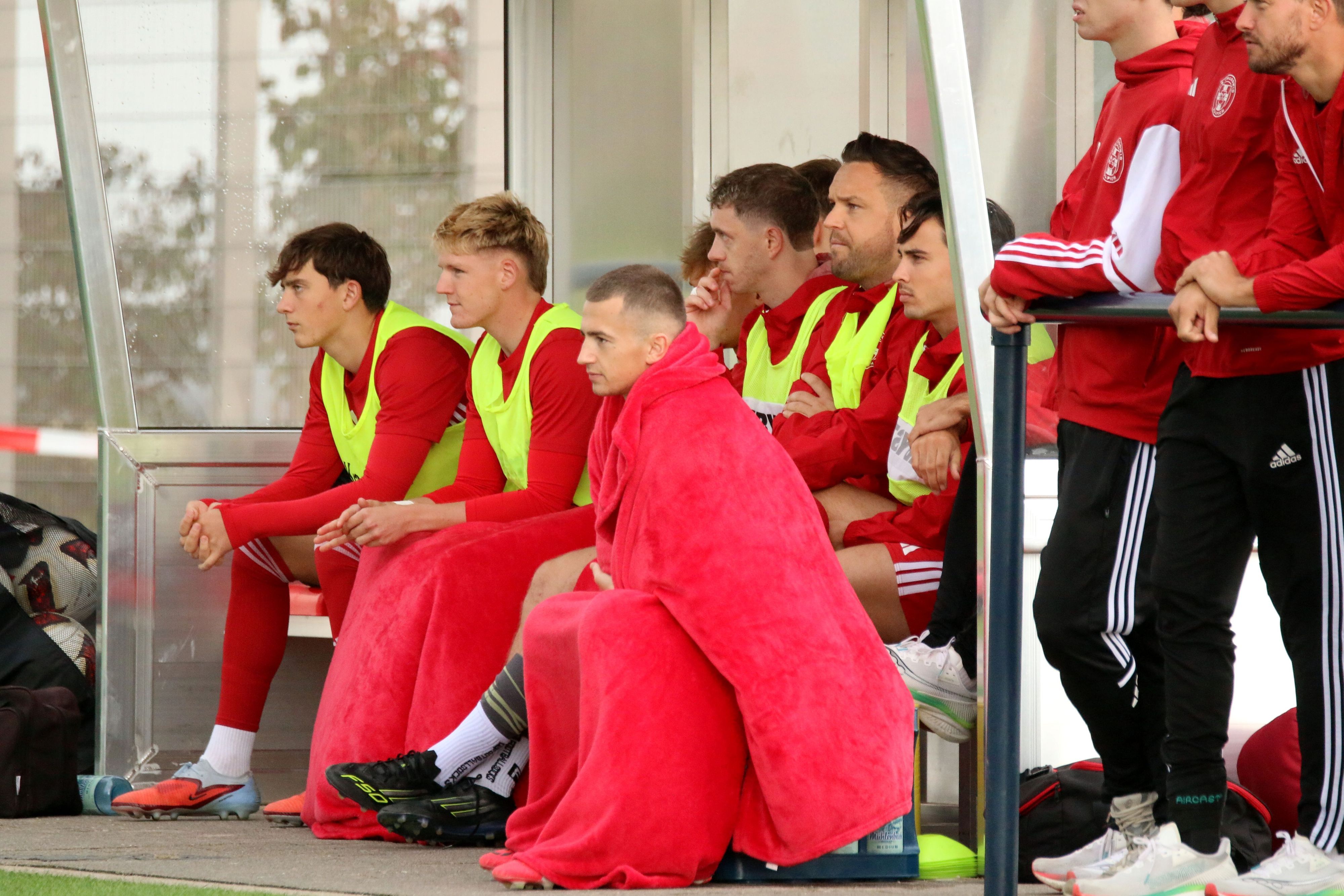 Ein Zülpicher Auswechelspieler hat sich komplett in rote Decken gehüllt, ein weiterer hat die Beine bedeckt.