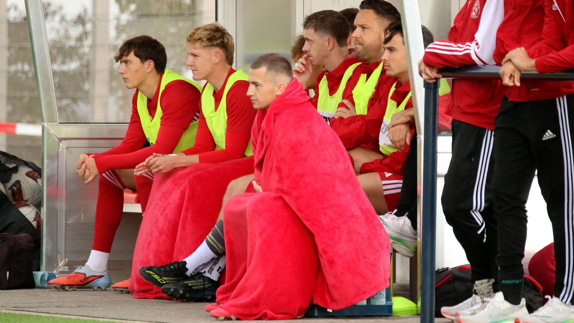 Ein Zülpicher Auswechelspieler hat sich komplett in rote Decken gehüllt, ein weiterer hat die Beine bedeckt.