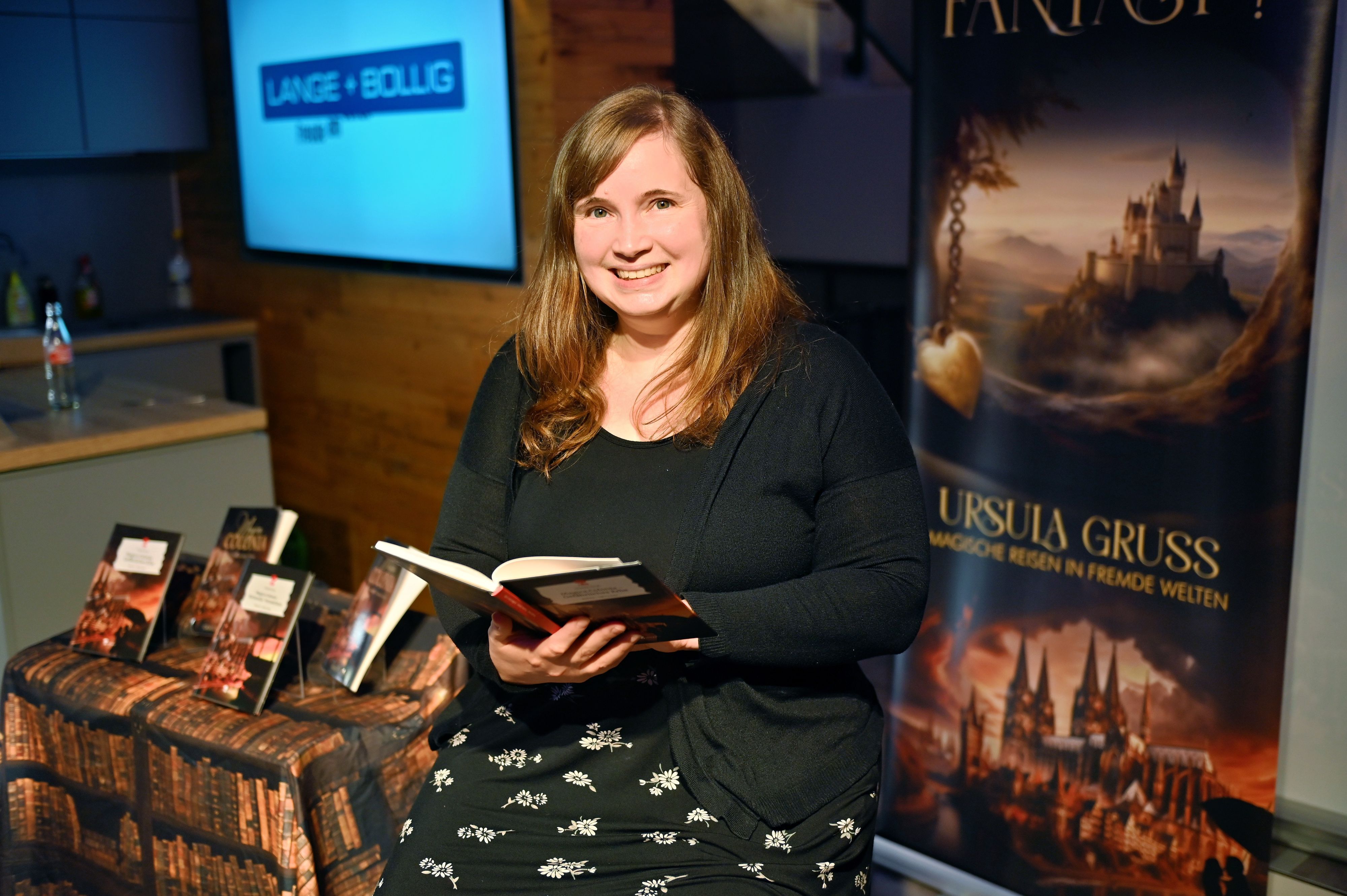 Autorin Ursula Gruß mit einem aufgeschlagenen Buch in der Hand vor einem Roll-up mit ihrem Namen.