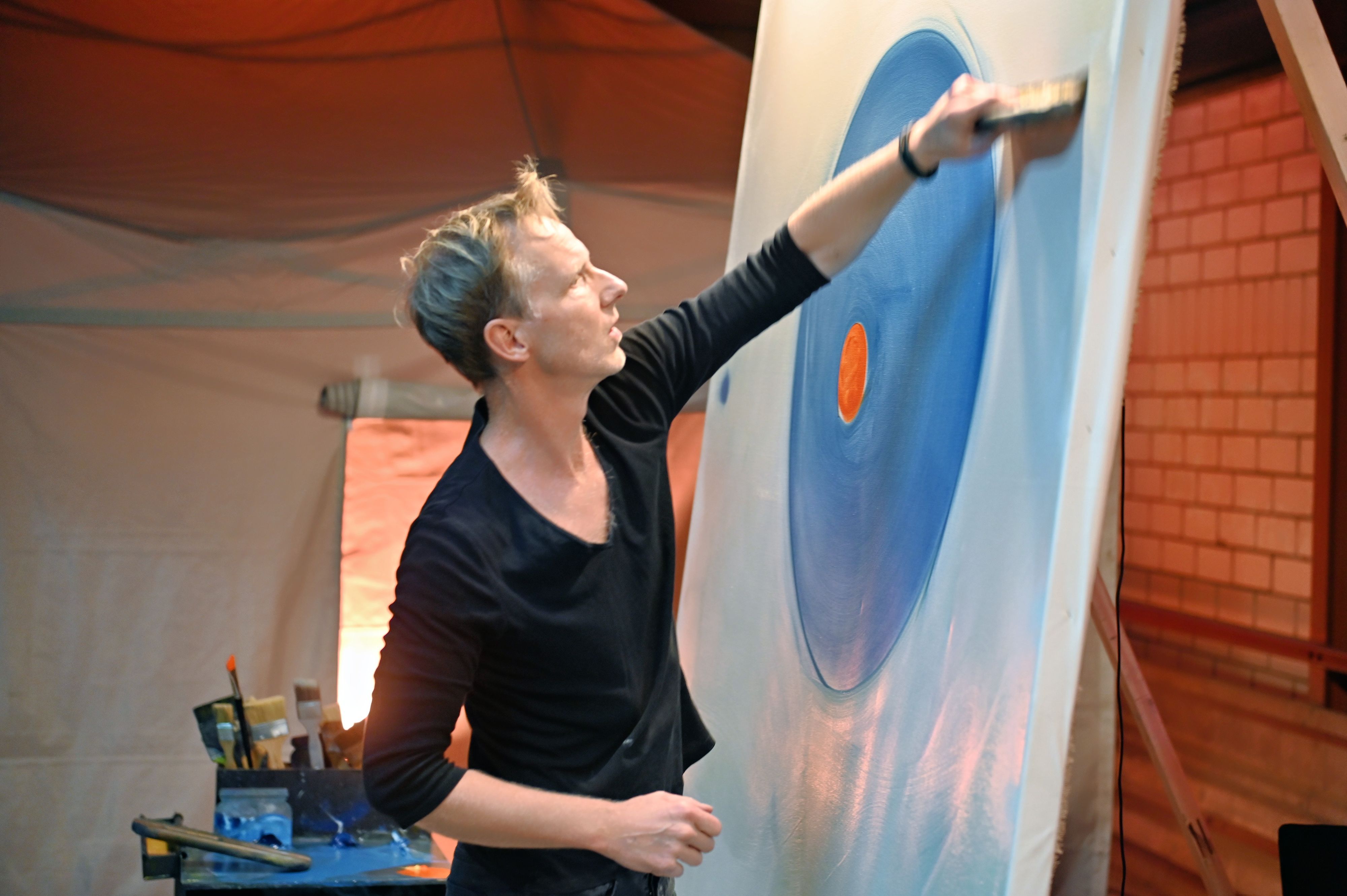 Maler Marc Westermann arbeitet an einem großen Bild. Auf der Leinwand sind ein blauer Farbkreis mit einem roten Punkt in der Mitte zu sehen.