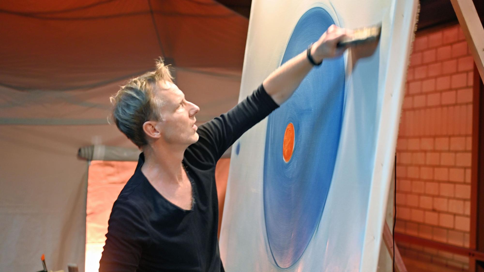 Maler Marc Westermann arbeitet an einem großen Bild. Auf der Leinwand sind ein blauer Farbkreis mit einem roten Punkt in der Mitte zu sehen.