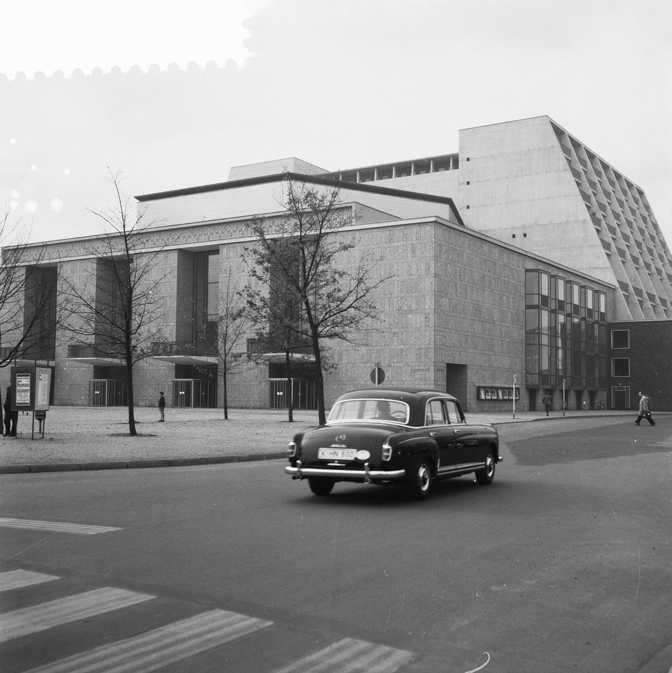 Auf einer alten Fotoaufnahme sieht man ein Auto an einem Gebäude vorbeifahren.
