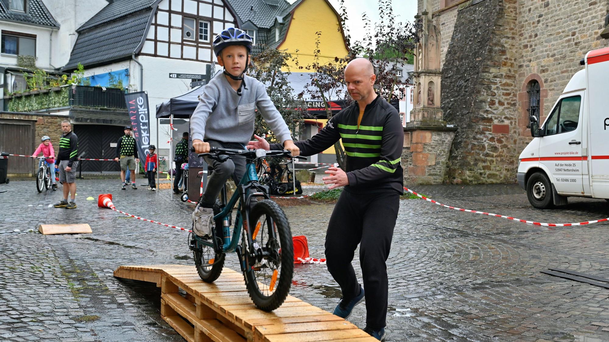Ein Junge fährt mit dem Fahrrad über eine hölzerne Rampe, ein Mann gibt Hilfestellung.