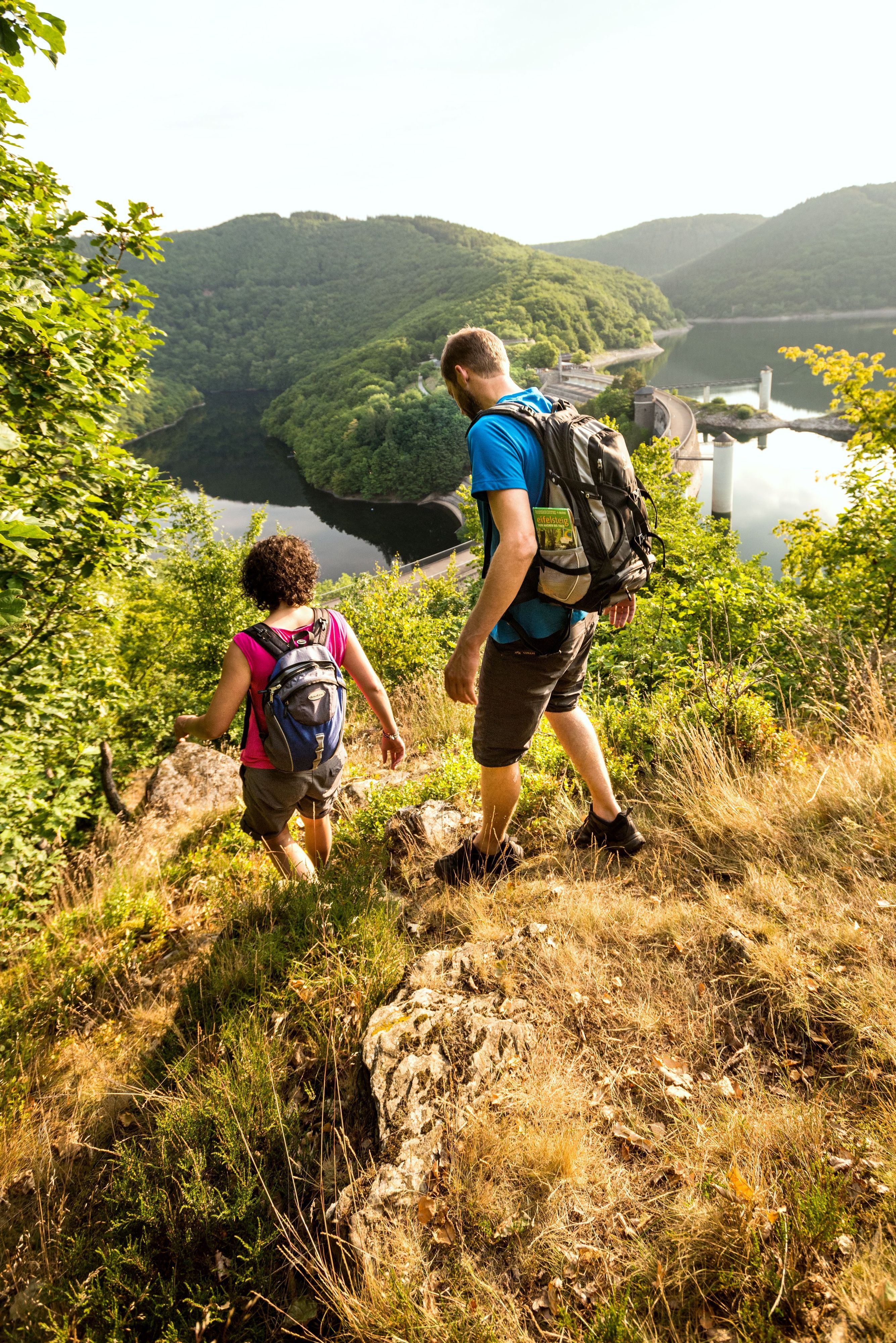 Zwei Wanderer mit Rucksäcken, die auf dem Foto nur mit dem Rücken zu sehen sind, steigen einen Weg in Richtung der Staumauer des Urftsees hinab.
