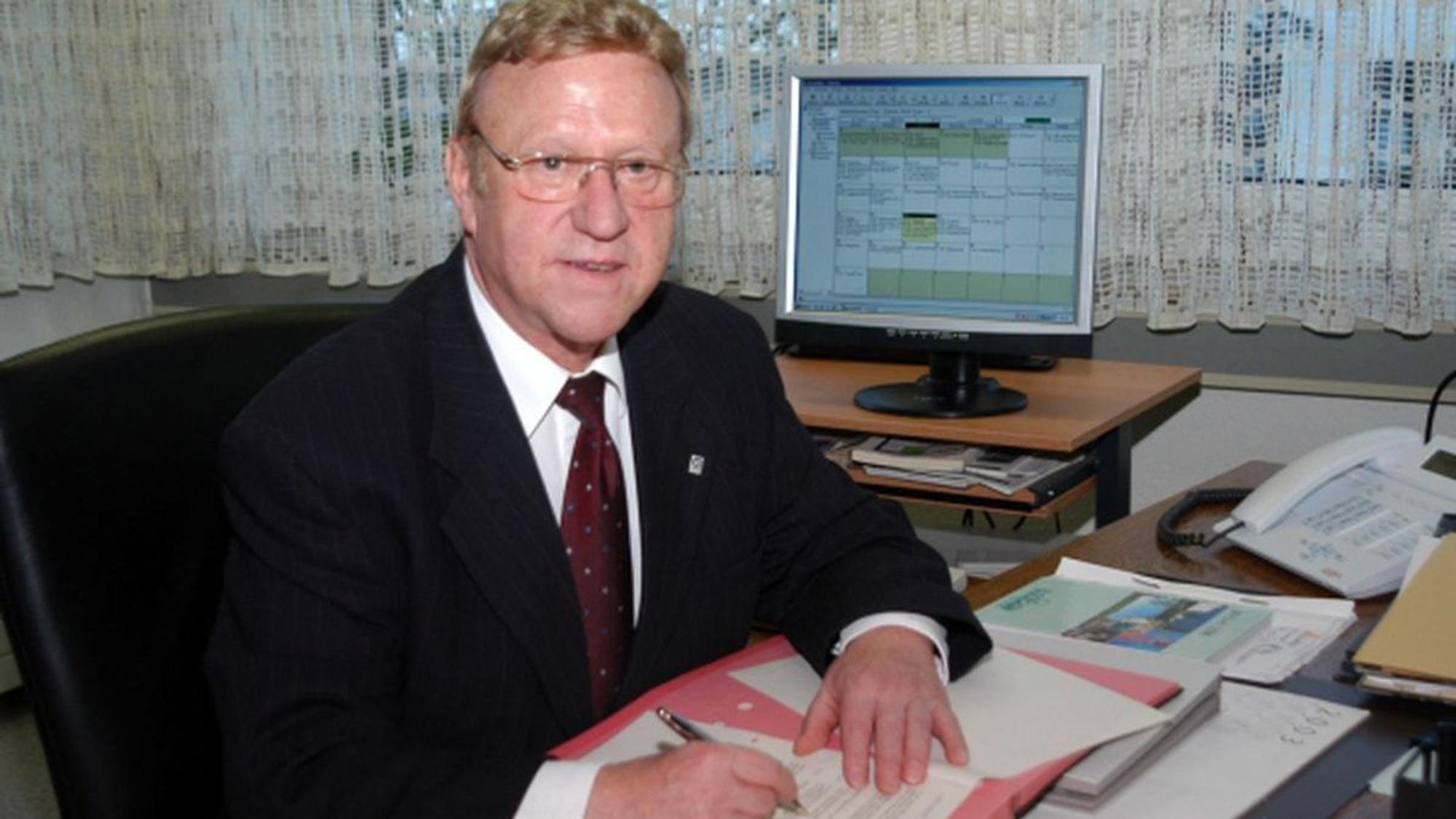 Hans Kaiser sitzt an seinem Schreibtisch, einen Stift und Unterlagen vor sich liegend.