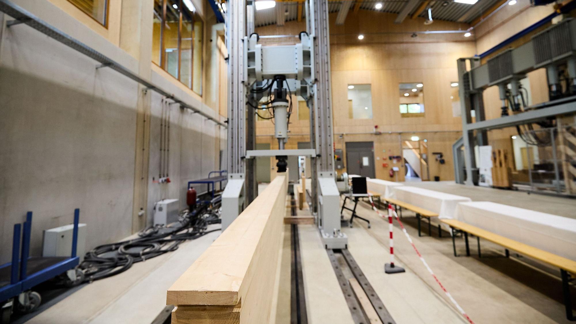 Ein langer Holzbalken ist in einer großen Werkhalle in ein Prüfgerät eingespannt.