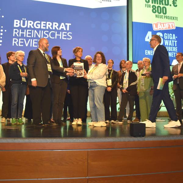Das Bild zeigt den Bürgerrat auf der Bühne. Vertreter übergeben Ministerin Mona Neubaur einen Katalog mit 100 Vorschlägen für den Strukturwandel.