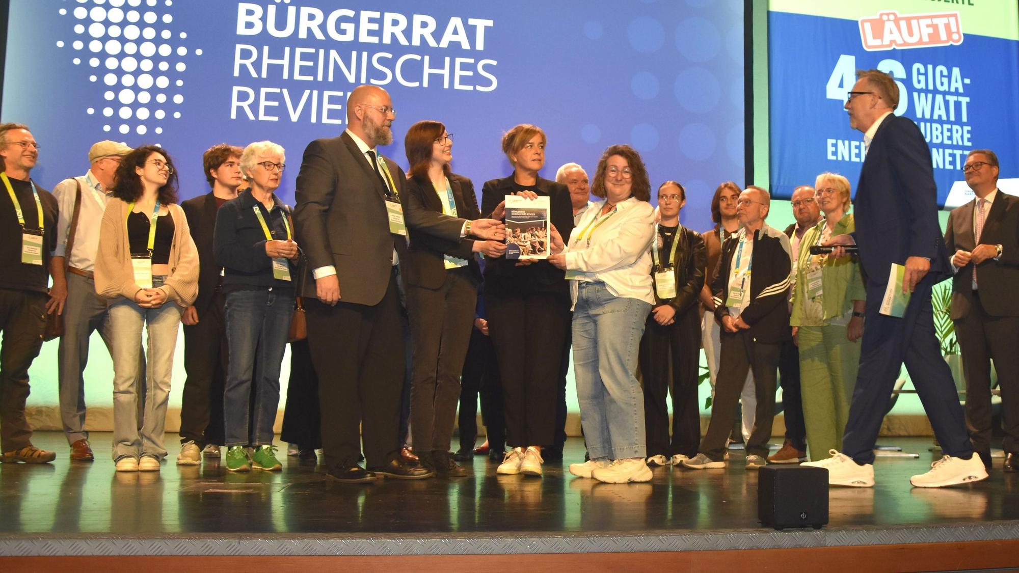 Das Bild zeigt den Bürgerrat auf der Bühne. Vertreter übergeben Ministerin Mona Neubaur einen Katalog mit 100 Vorschlägen für den Strukturwandel.