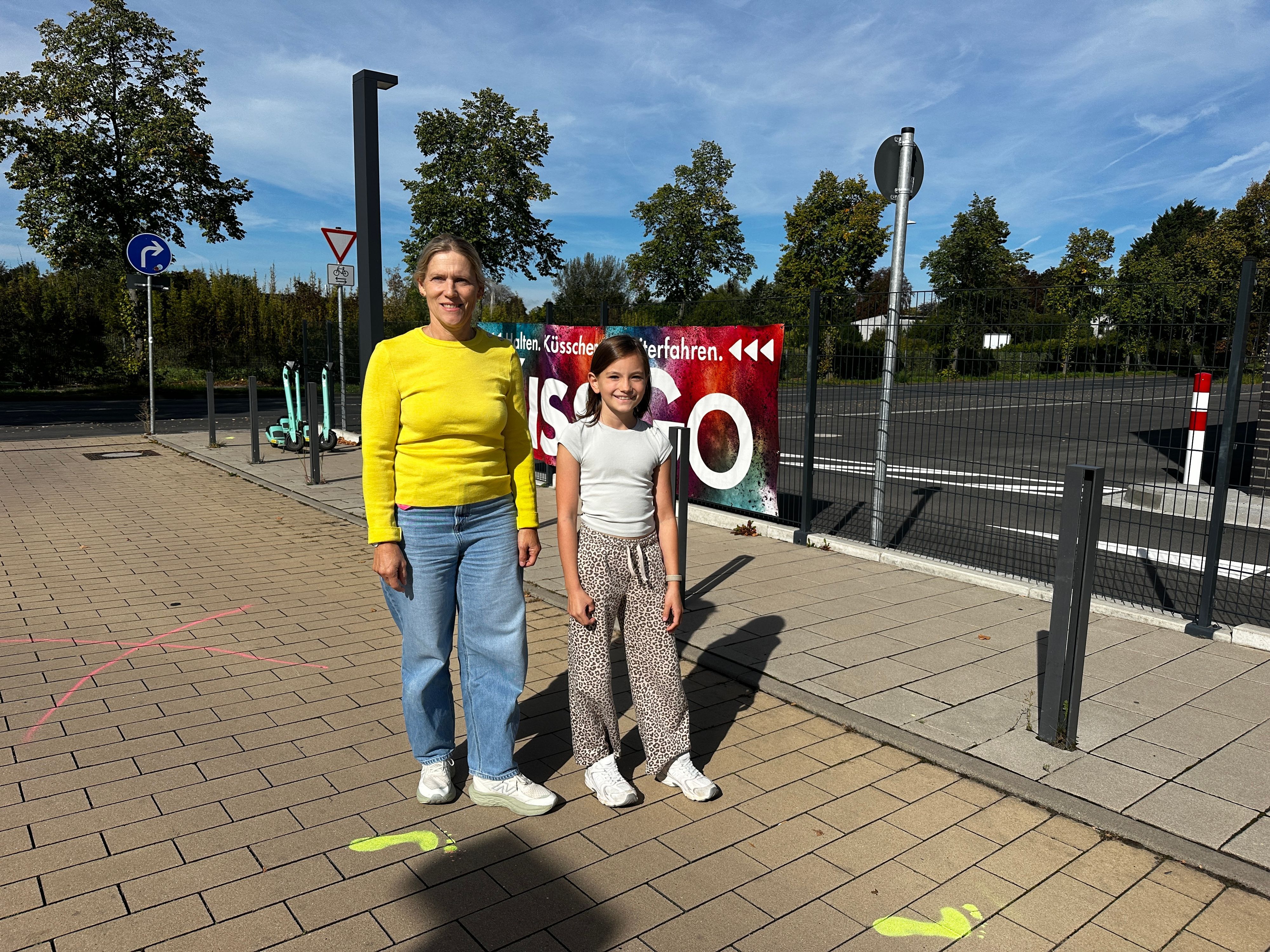 Eine Frau und ein Mädchen stehen auf einem gepflasterten Weg, auf dem farbige Fußabdrücke aufgesprüht sind; im Hintergrund sind eine Straße, Verkehrsschilder, Bäume und ein Zaun zu sehen.