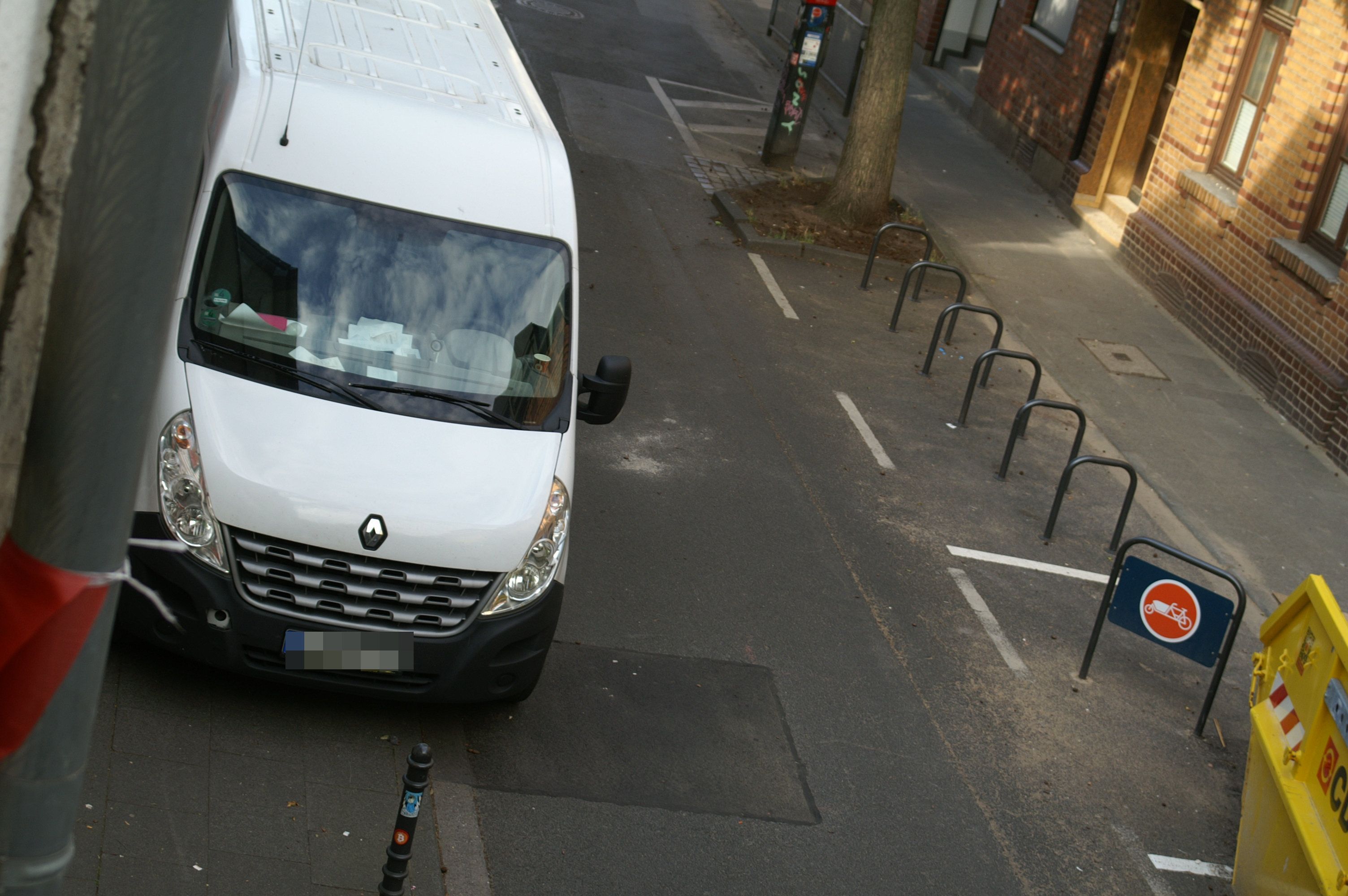 Ein weißer Transporter parkt in einer schmalen Straße gegenüber einem Fahrradparkplatz.