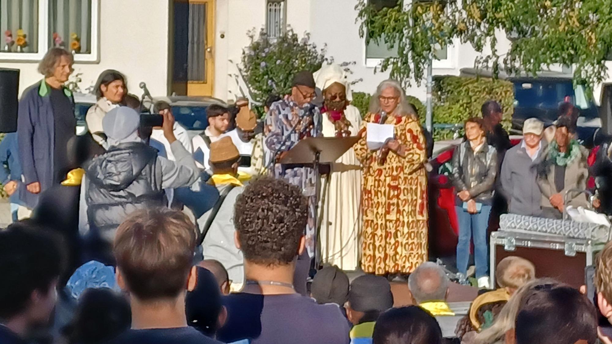 Viele Menschen stehen auf der Straße und hören drei in afrikanische Gewänder gekleideten Personen zu, die in Mikrofone sprechen.