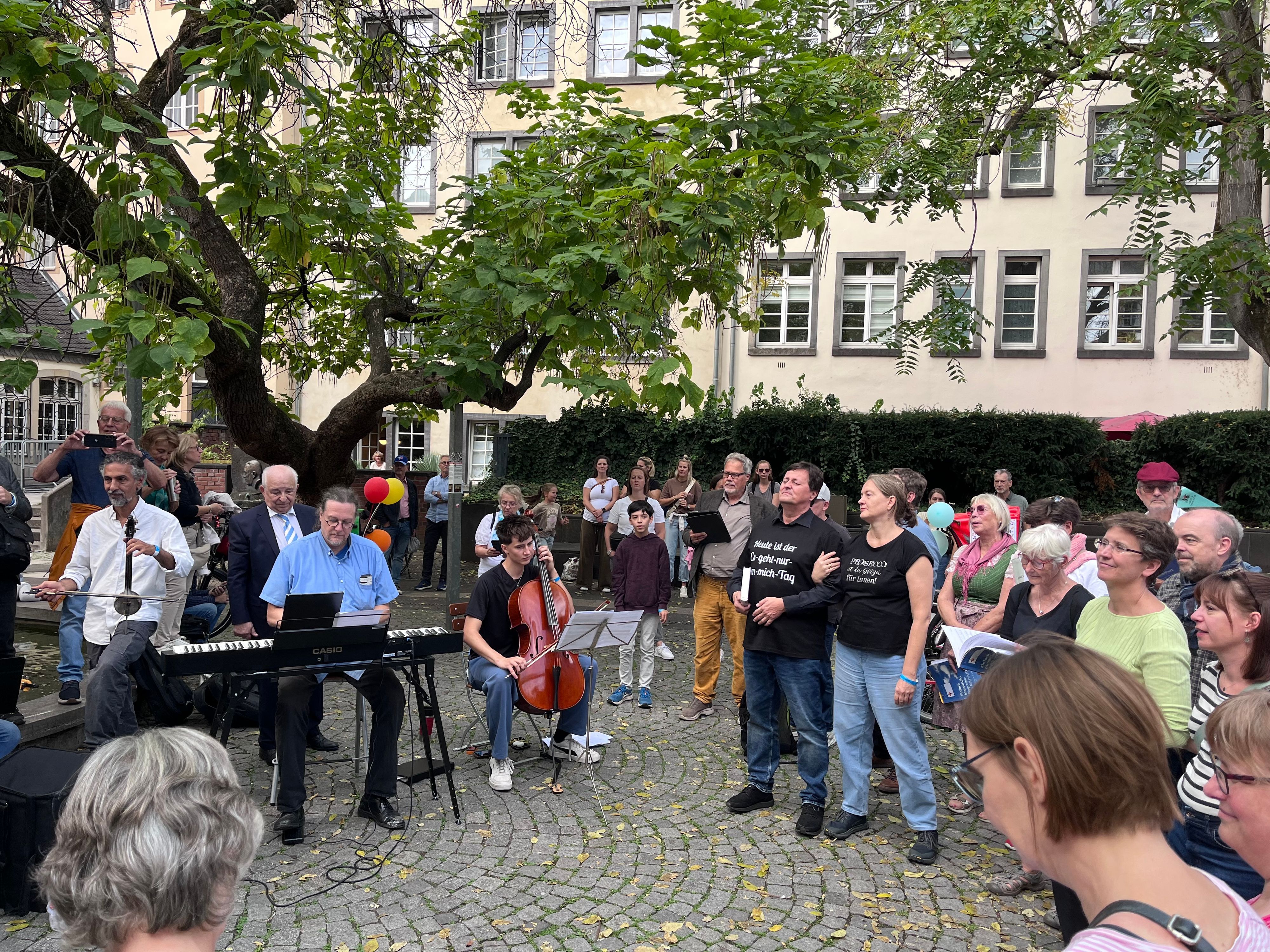 Viele Menschen stehen im Halbkreis. Unter einem alten Baum musizieren Musiker mit Keyboard, Cello und einem orientalischen Instrument.