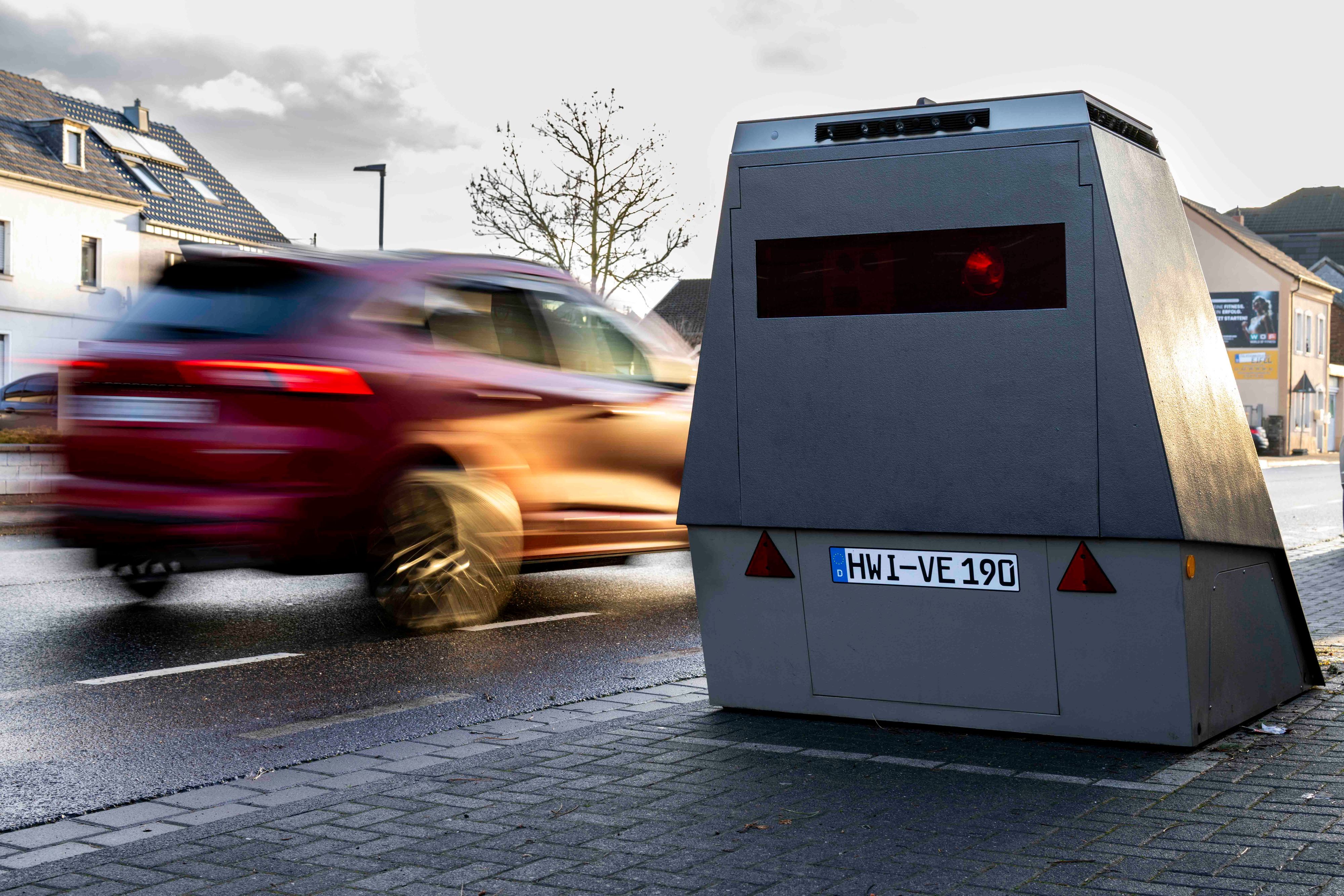 Ein rotes Auto fährt an einem Blitzeranhänger vorbei, der am Straßenrand abgestellt ist.