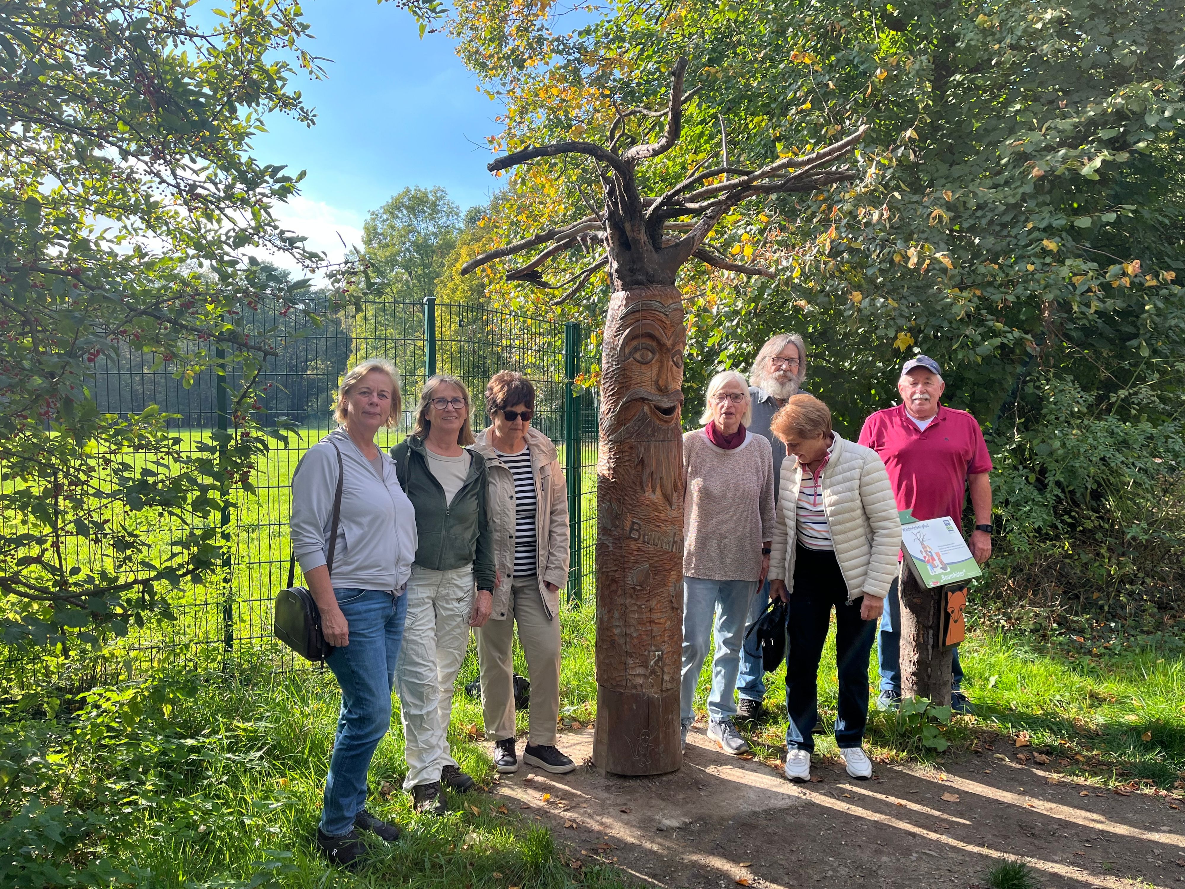 Sieben Erwachsene stehen neben einer hohen, geschnitzten Holzfigur mit Gesicht und ausladenden Ästen. Im Hintergrund sind ein Zaun und ein grünes Gehege zu sehen.