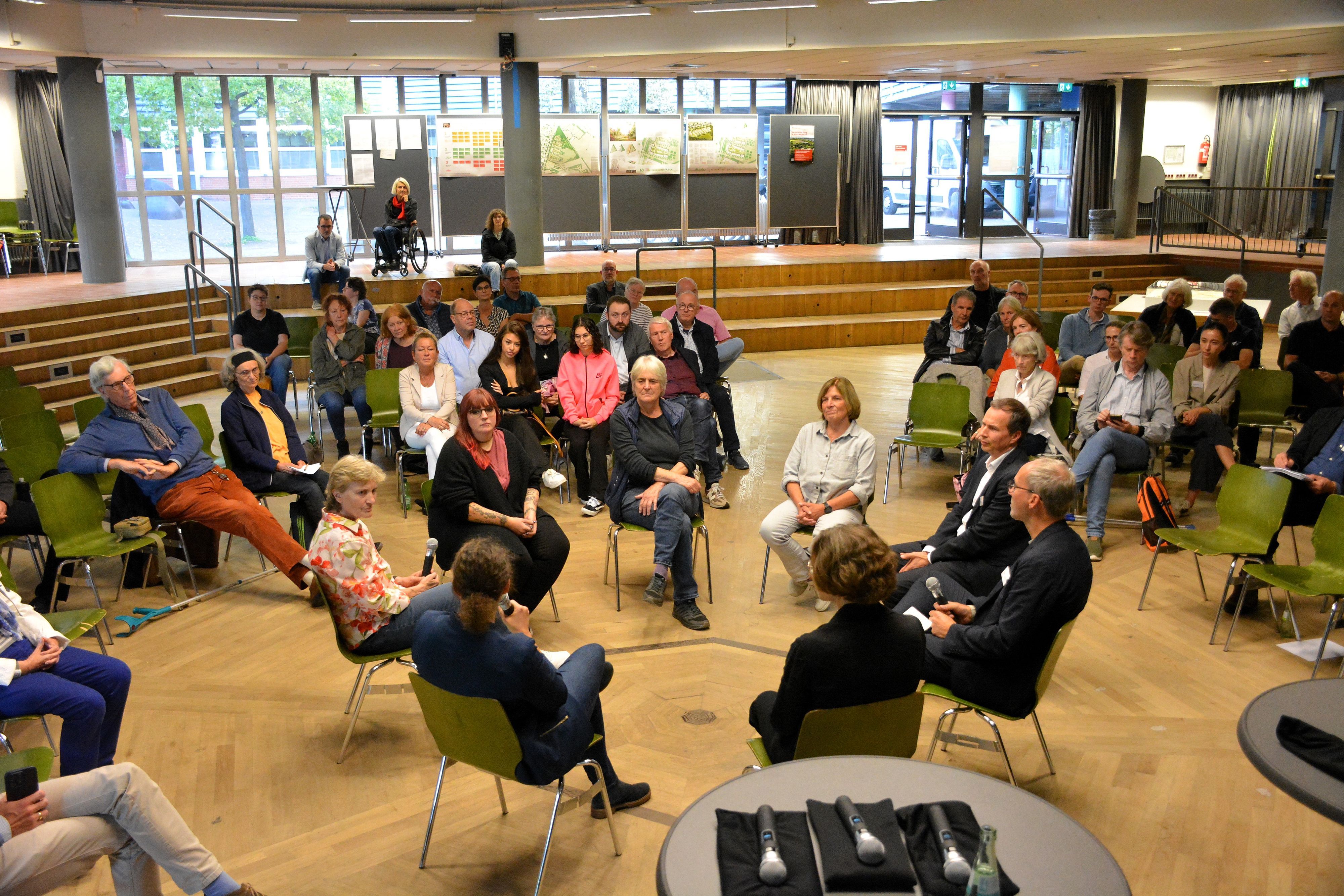Acht Leute sitzen in einem Stuhlkreis und diskutieren vor Publikum.