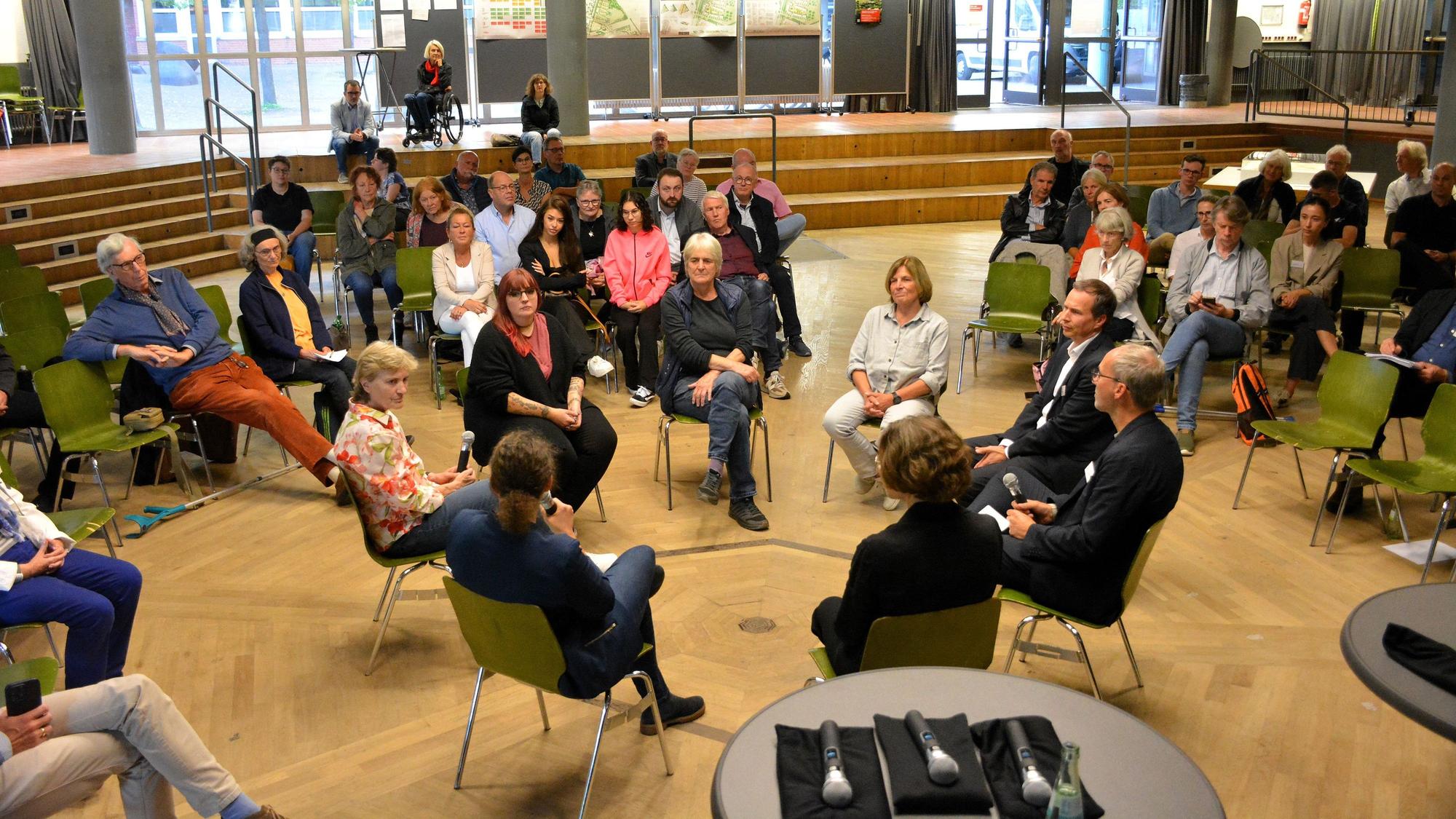 Acht Leute sitzen in einem Stuhlkreis und diskutieren vor Publikum.