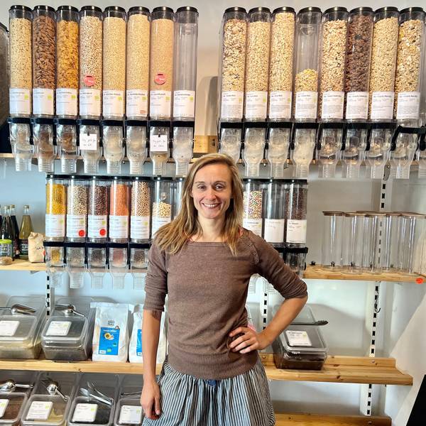 Eine Frau mit langem, mittelblondem Haar steht in einem Ladenlokal vor einem Regal, in dem gefüllte Unverpackt-Behälter zum Selbstabfüllen stehen.
