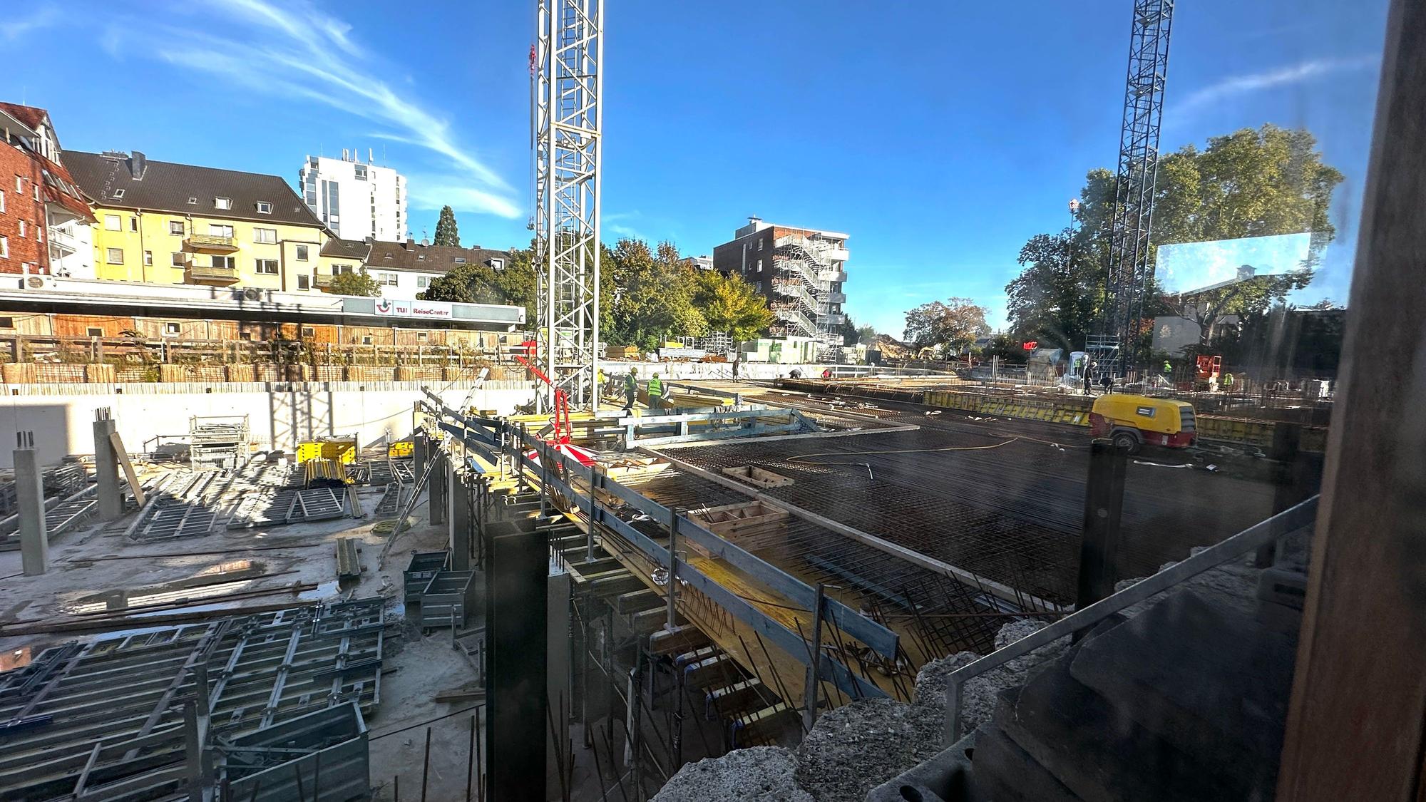 Zu sehen ist eine große Baustelle mit zwei Baukränen, einem teilweise betonierten Fundament und Stahlkonstruktionen, umgeben von Wohnhäusern und Bäumen.