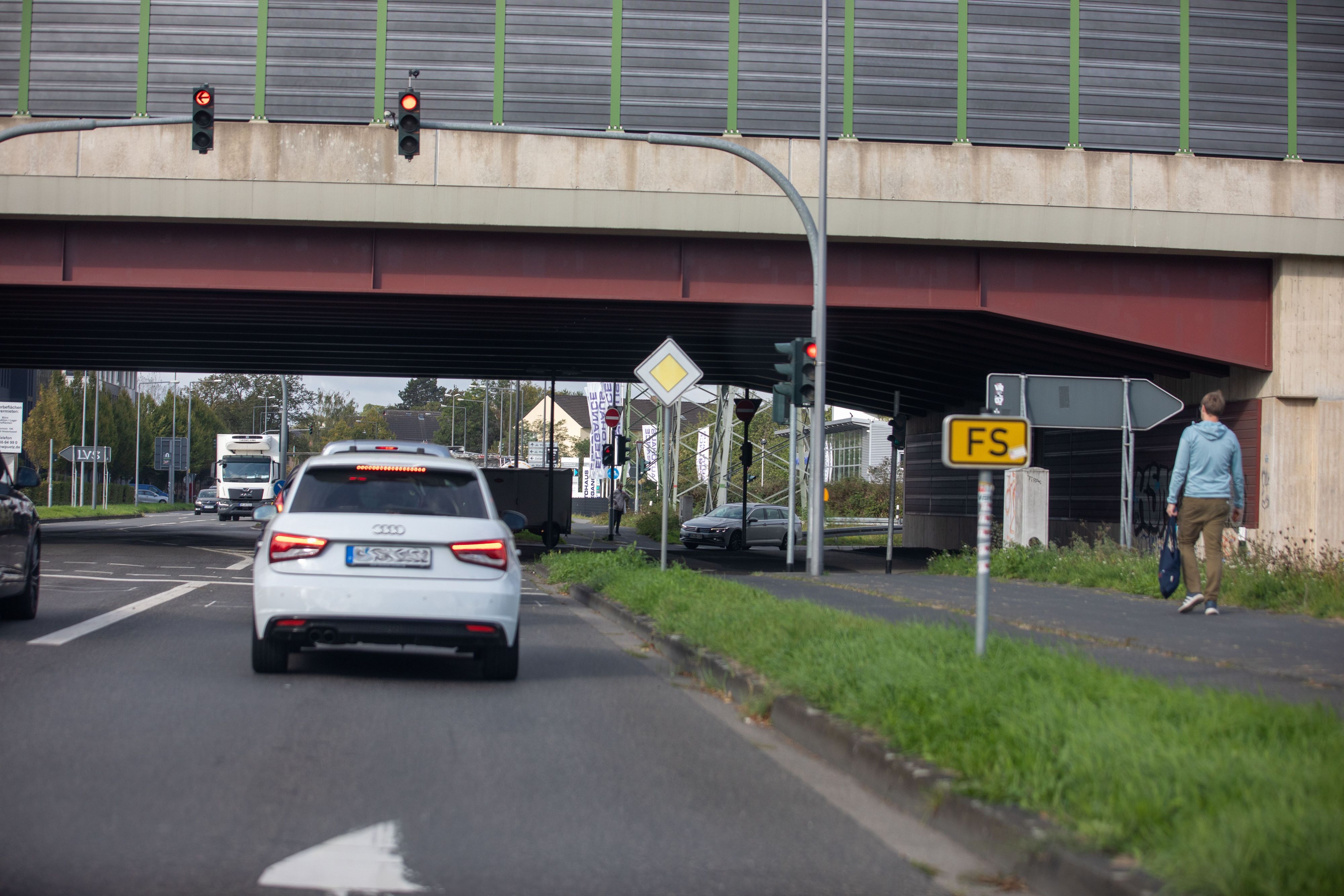 Eine stark befahrene Straßenkreuzung mit mehreren Fahrzeugen, Ampeln, Verkehrsschildern, Fahrbahnmarkierungen und einem Fußgänger neben den Straßen.